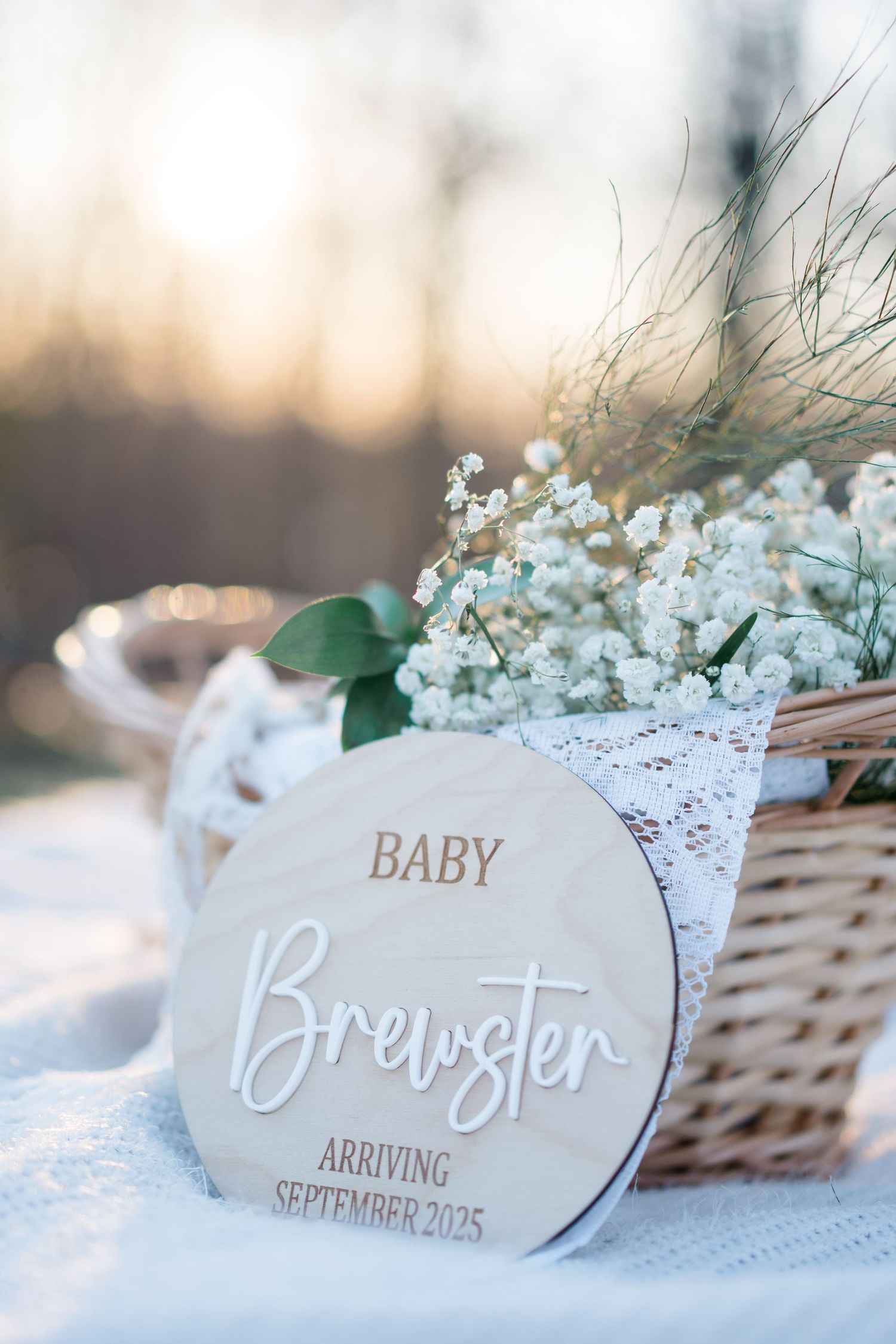 A wooden pregnancy announcement sign for Baby Brewster arriving September 2023 with baby's breath flowers and lace.