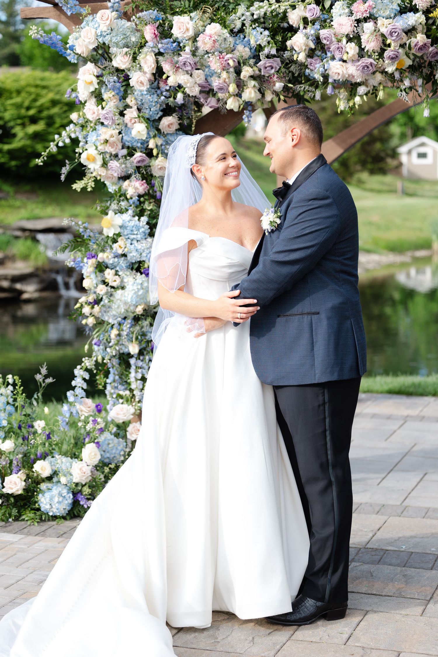 Wedding couple standing under floral arch with blue and white blooms near garden pond. Bear Brook Valley