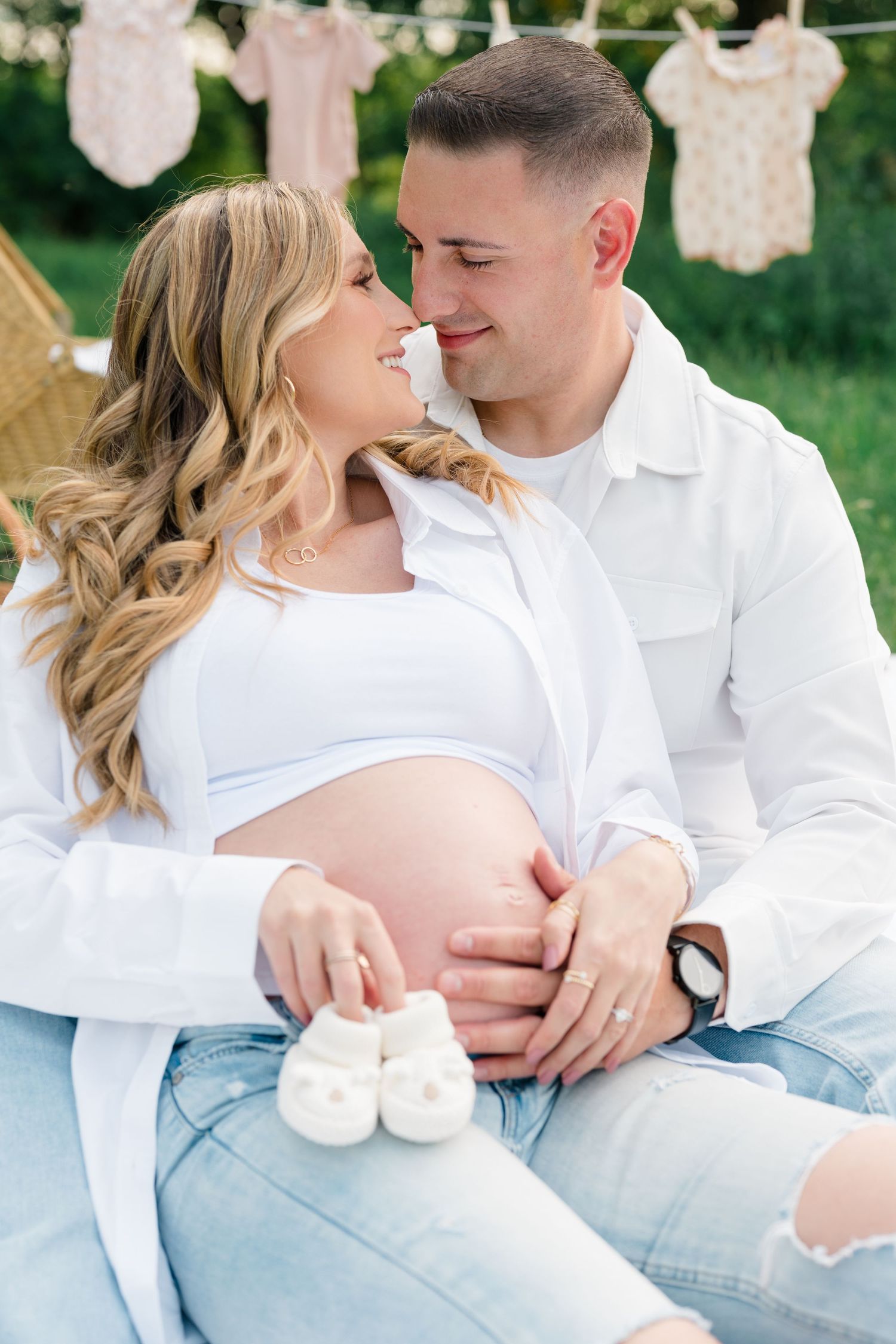 Intimate maternity photo with white outfits and tiny baby shoes in foreground against a garden backdrop.