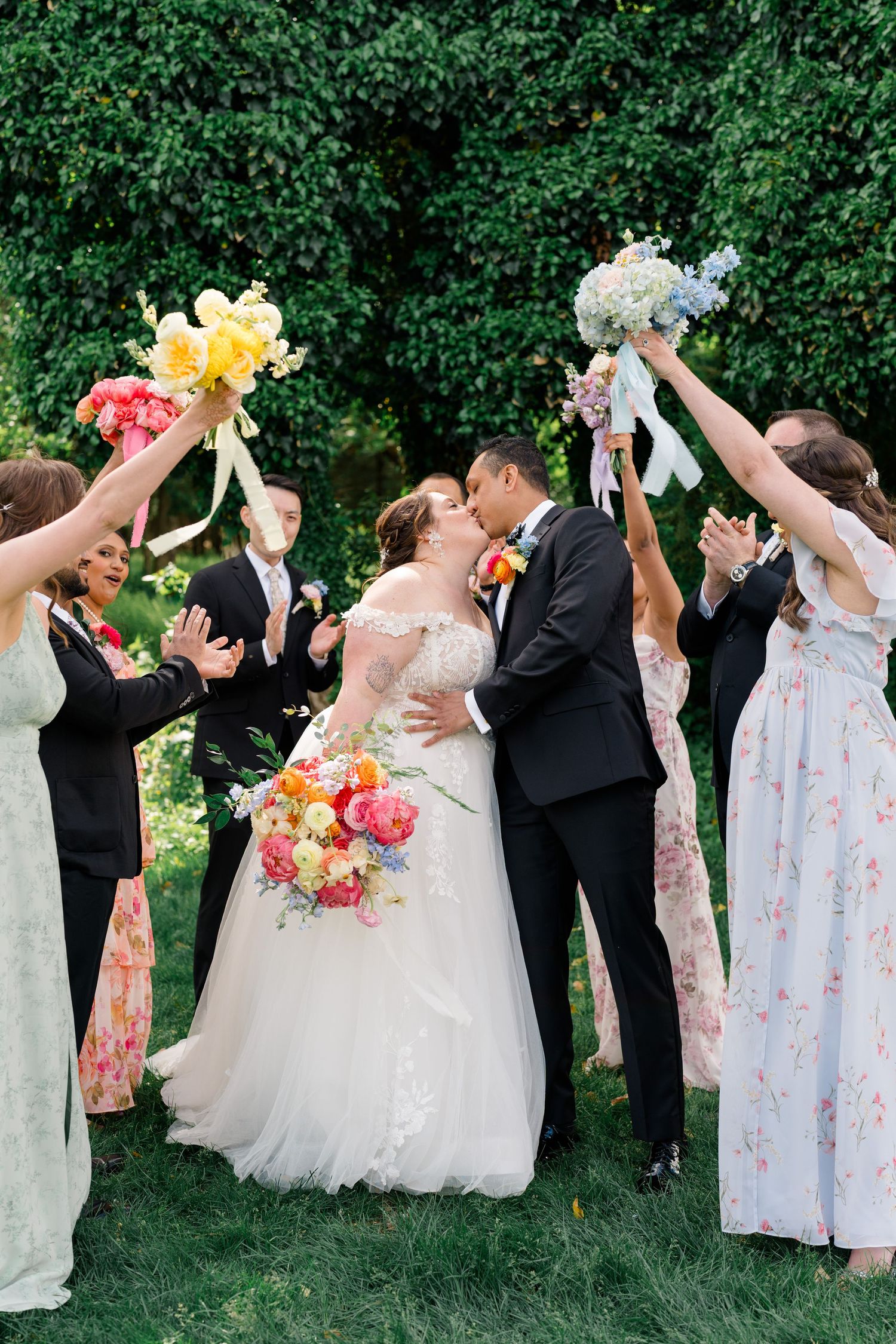 Wedding party celebrates with bouquets raised in the air against lush greenery backdrop.