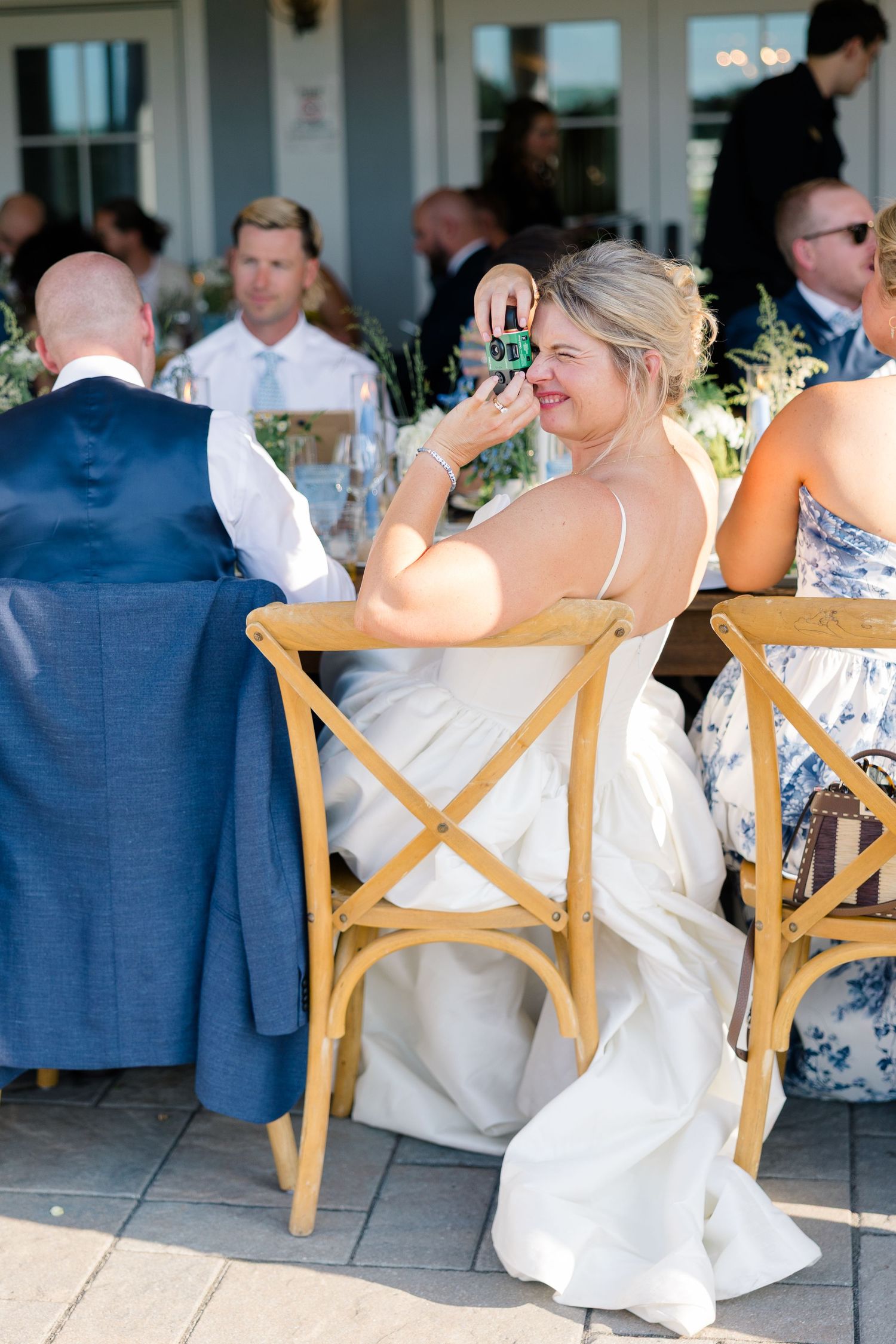 Smiling bride in white gown seated at reception table raising a small bottle in celebration.
