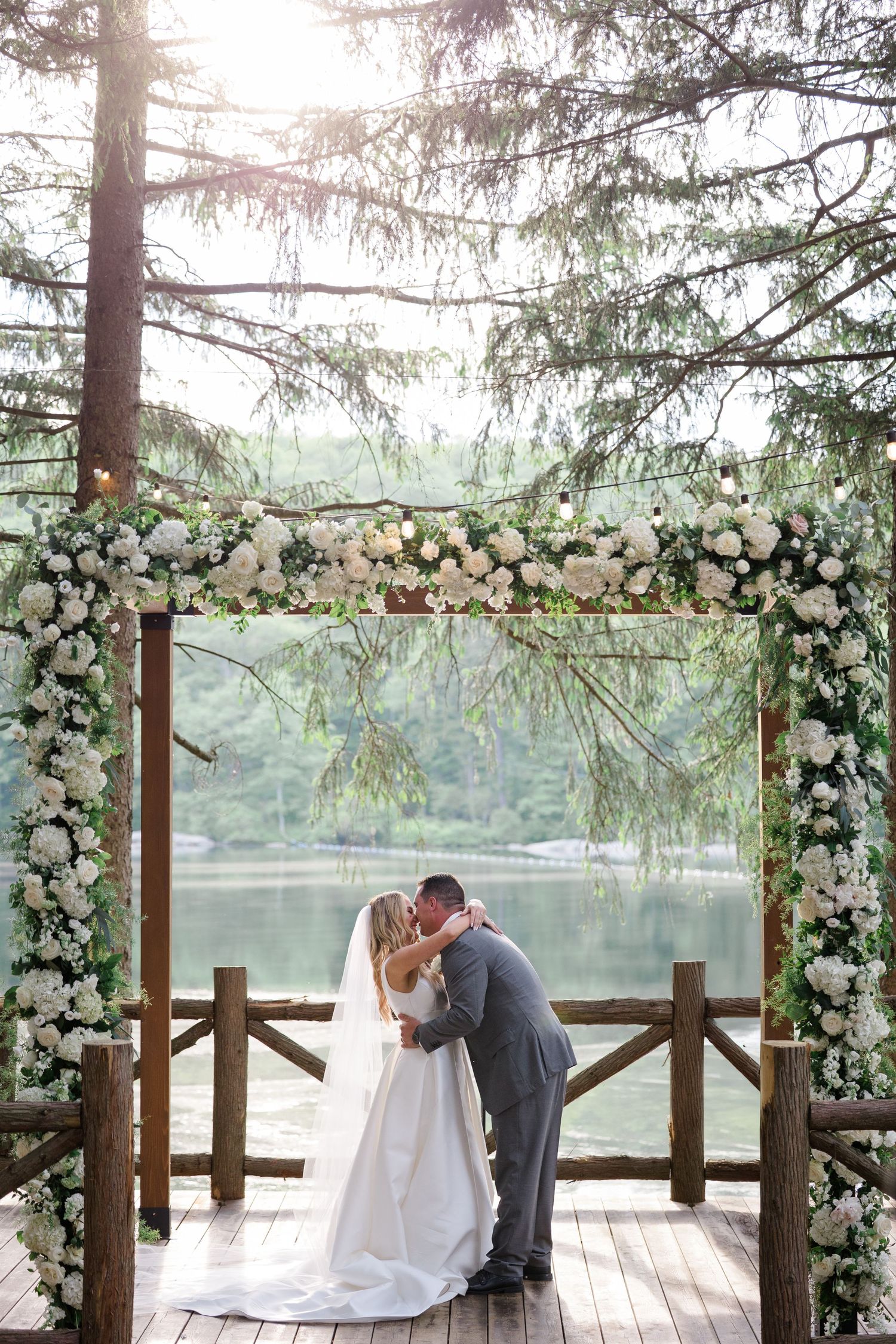 Wedding couple sharing a kiss under a floral arch on a wooden deck overlooking a lake.