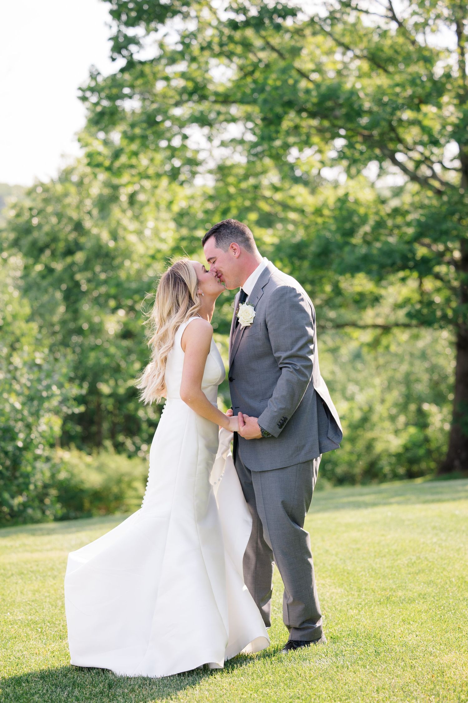 A newlywed couple embraces on a sunlit lawn with lush green trees in the background.