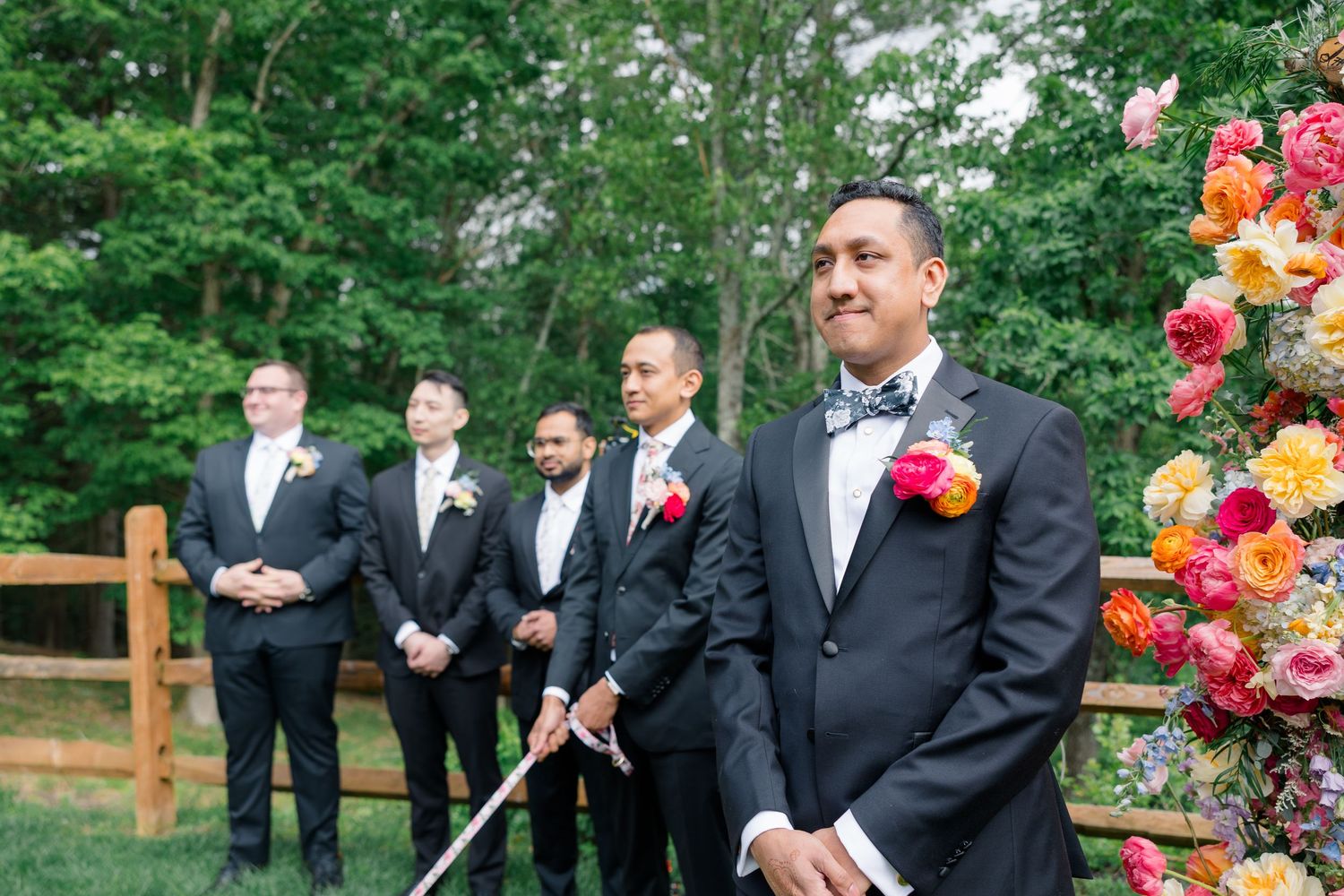 Groomsmen in black tuxedos standing at floral-decorated outdoor ceremony site.