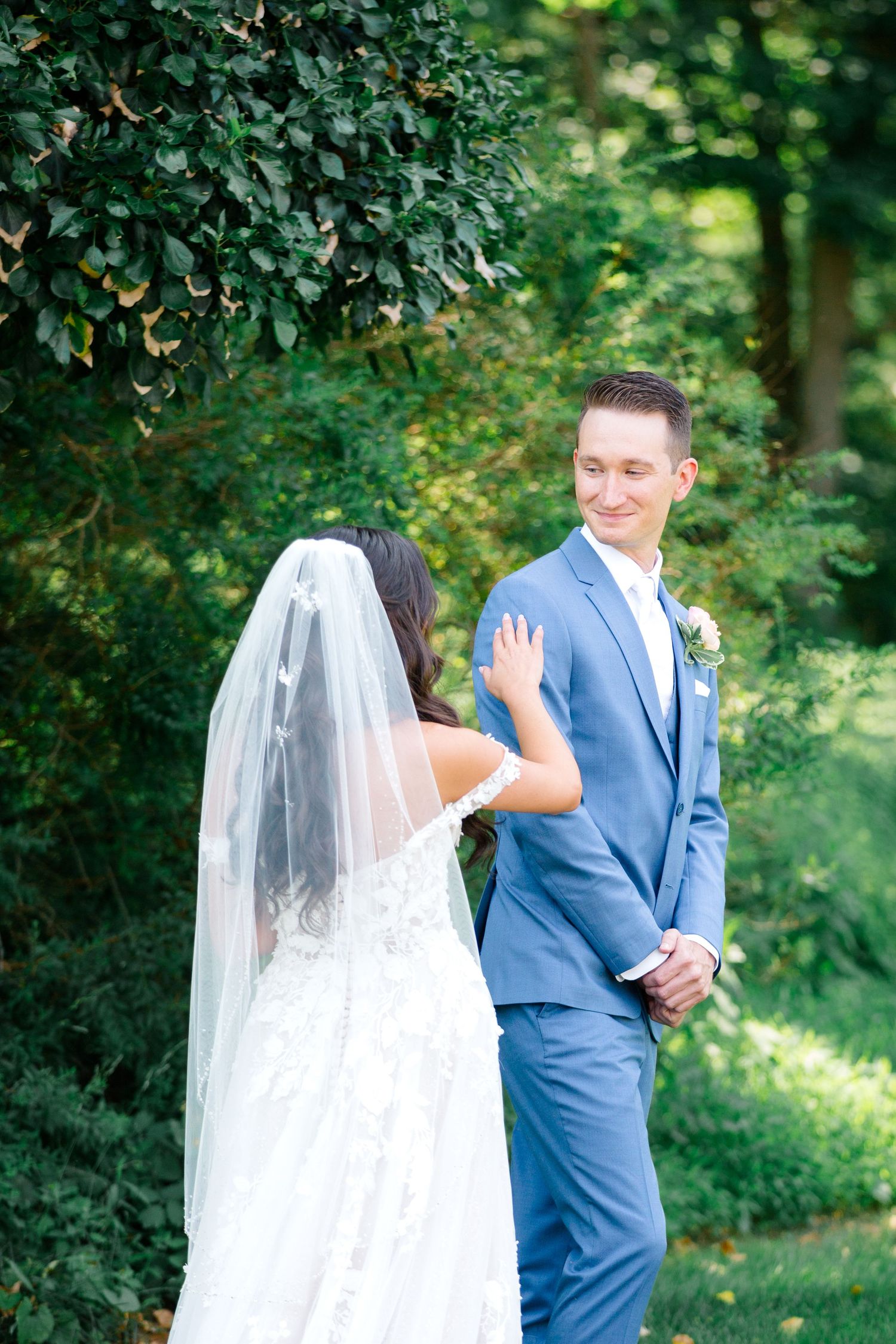 First look moment between bride and groom under green trees with bride wearing white veil.