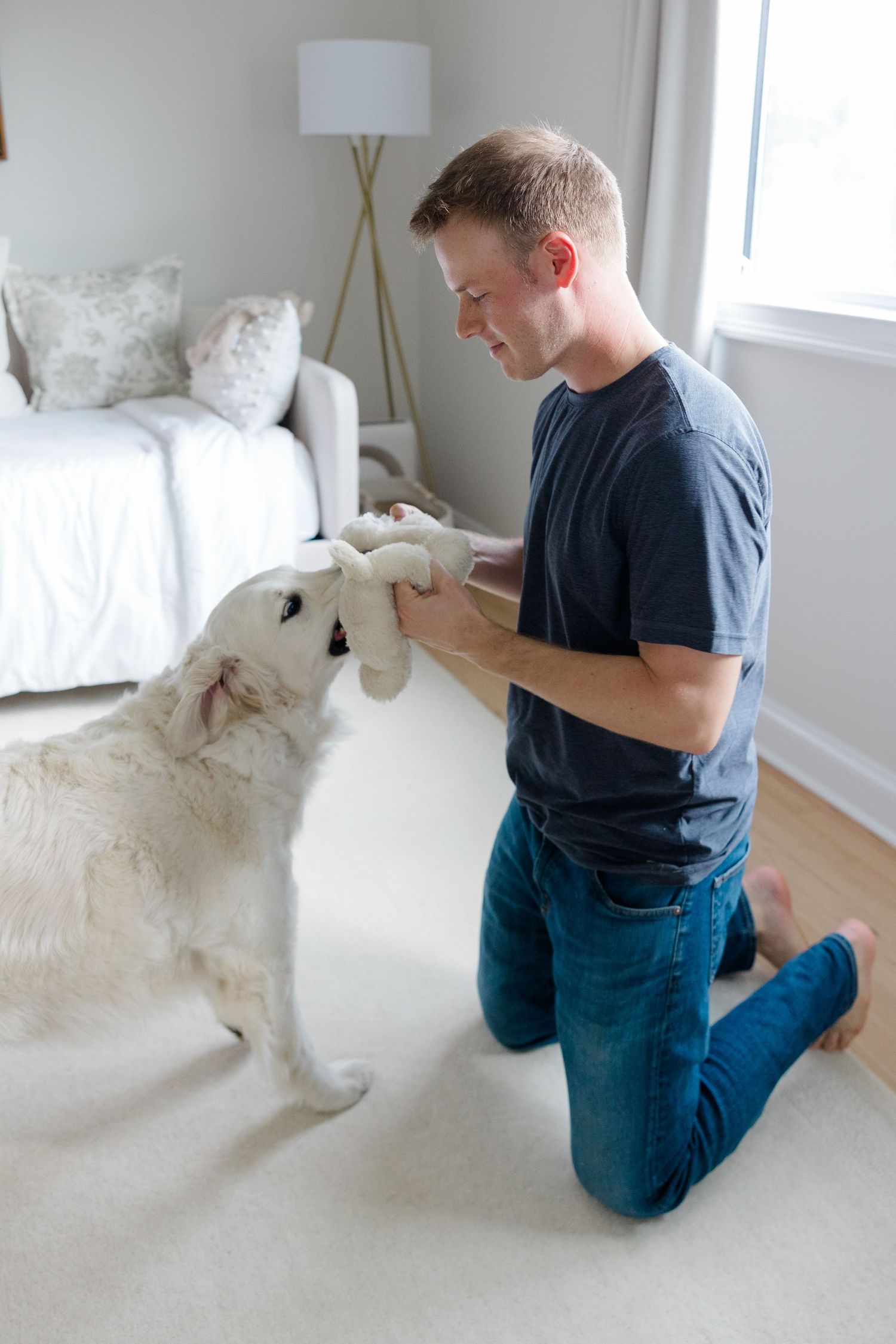 Person in blue shirt and jeans sits with white retriever dog on carpet while looking at phone in bright room.
