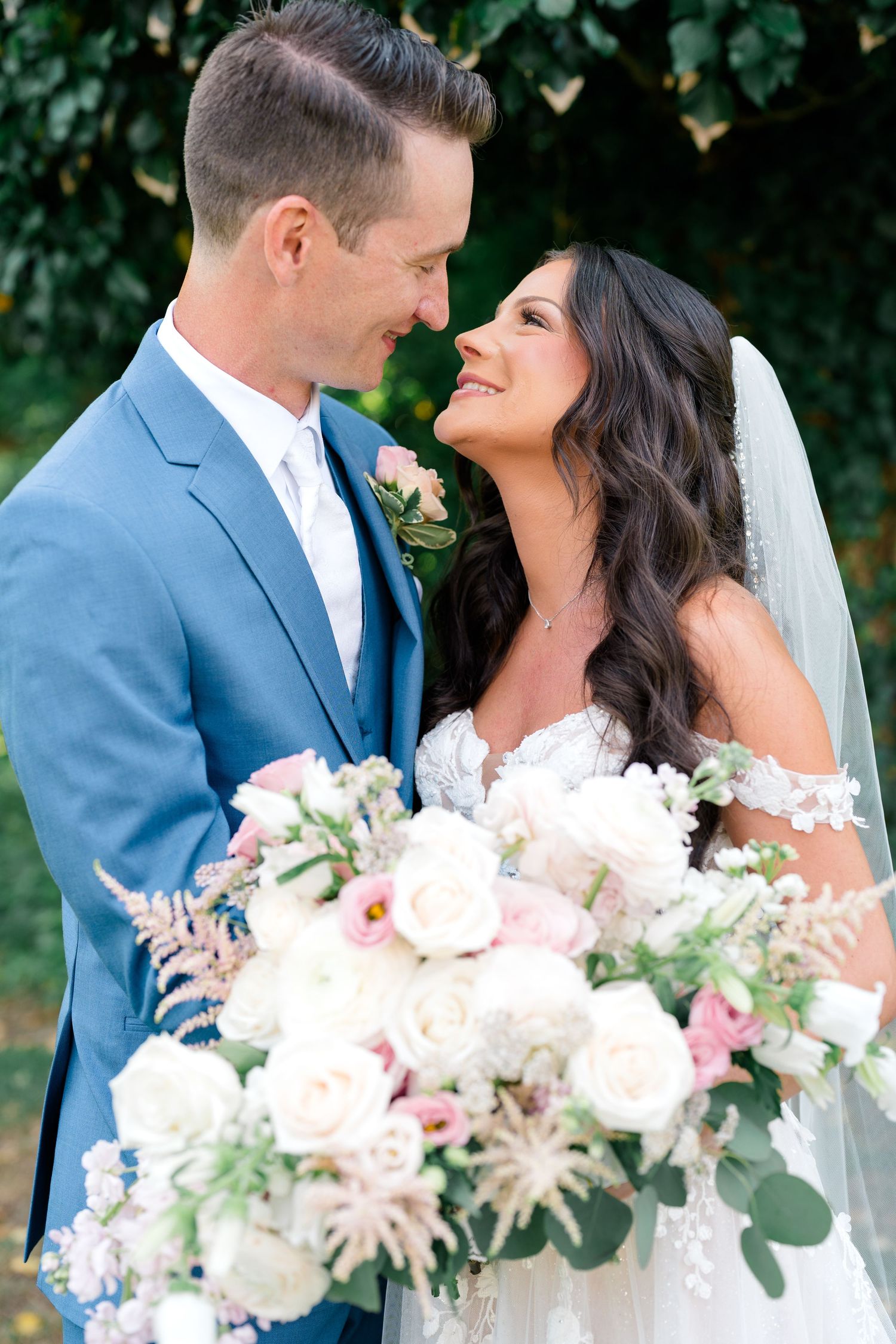 Bride and groom gazing lovingly at each other, bride holding lush white and pink floral bouquet, groom in blue suit.