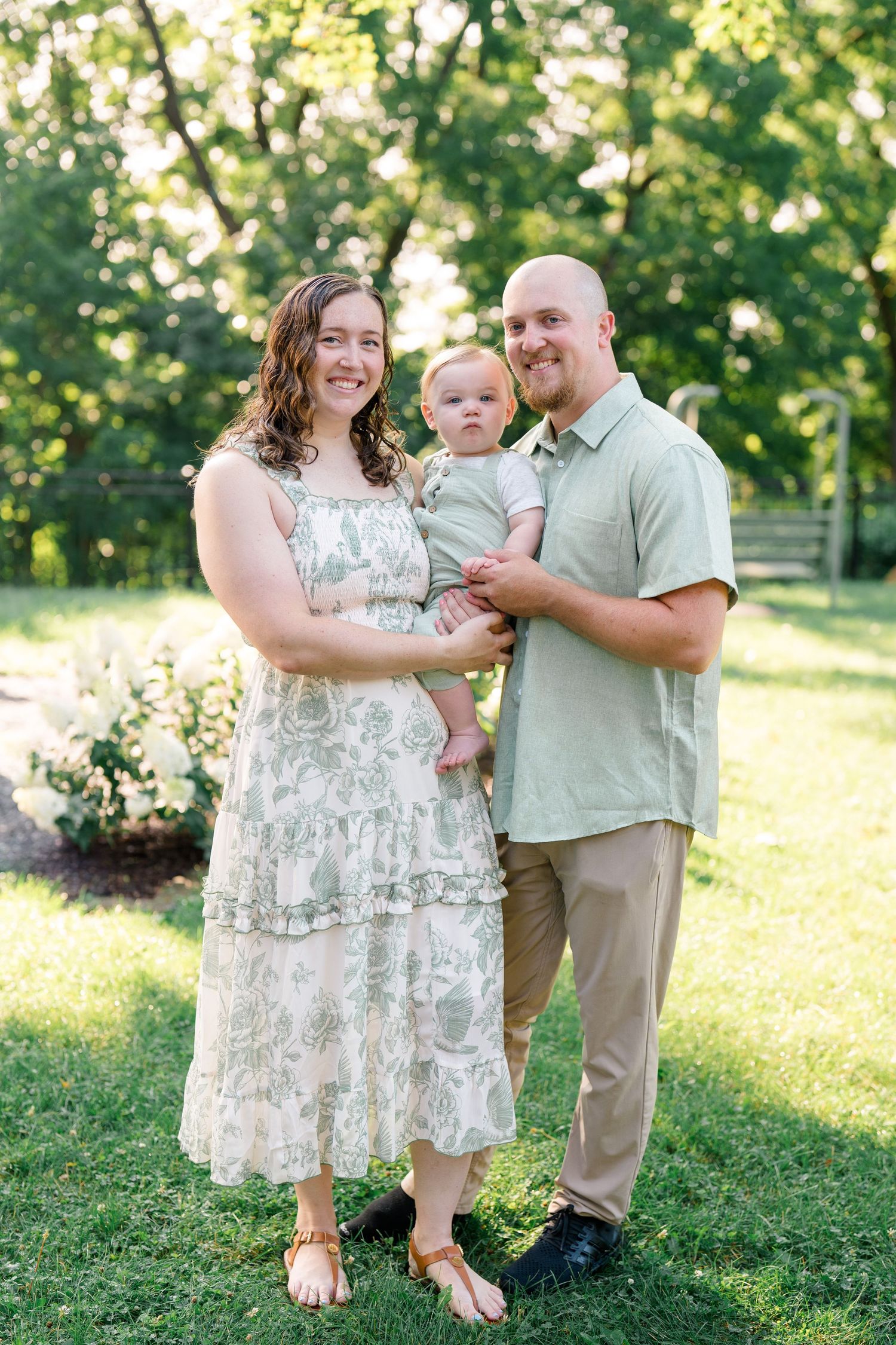 A smiling family of three poses outdoors, parents in sage green and floral dress holding their baby in a green romper.