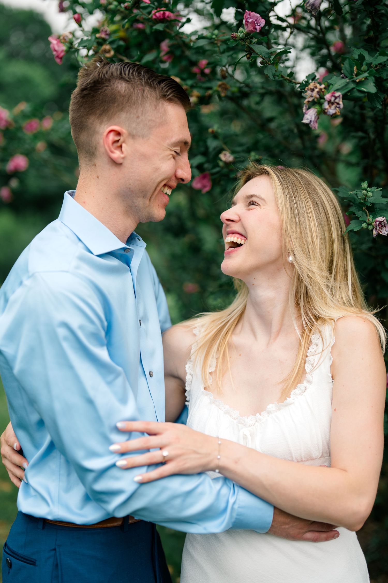 Laughing couple embracing outdoors near blooming flowers, man in blue shirt and woman in white dress.