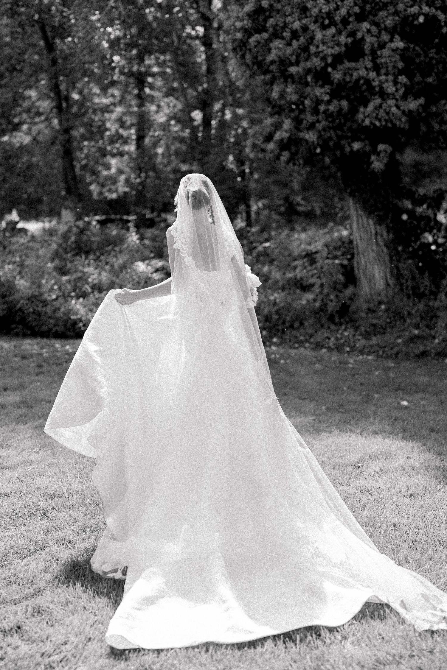 Black and white photo of a bride in a flowing white gown and veil walking through a lush garden.