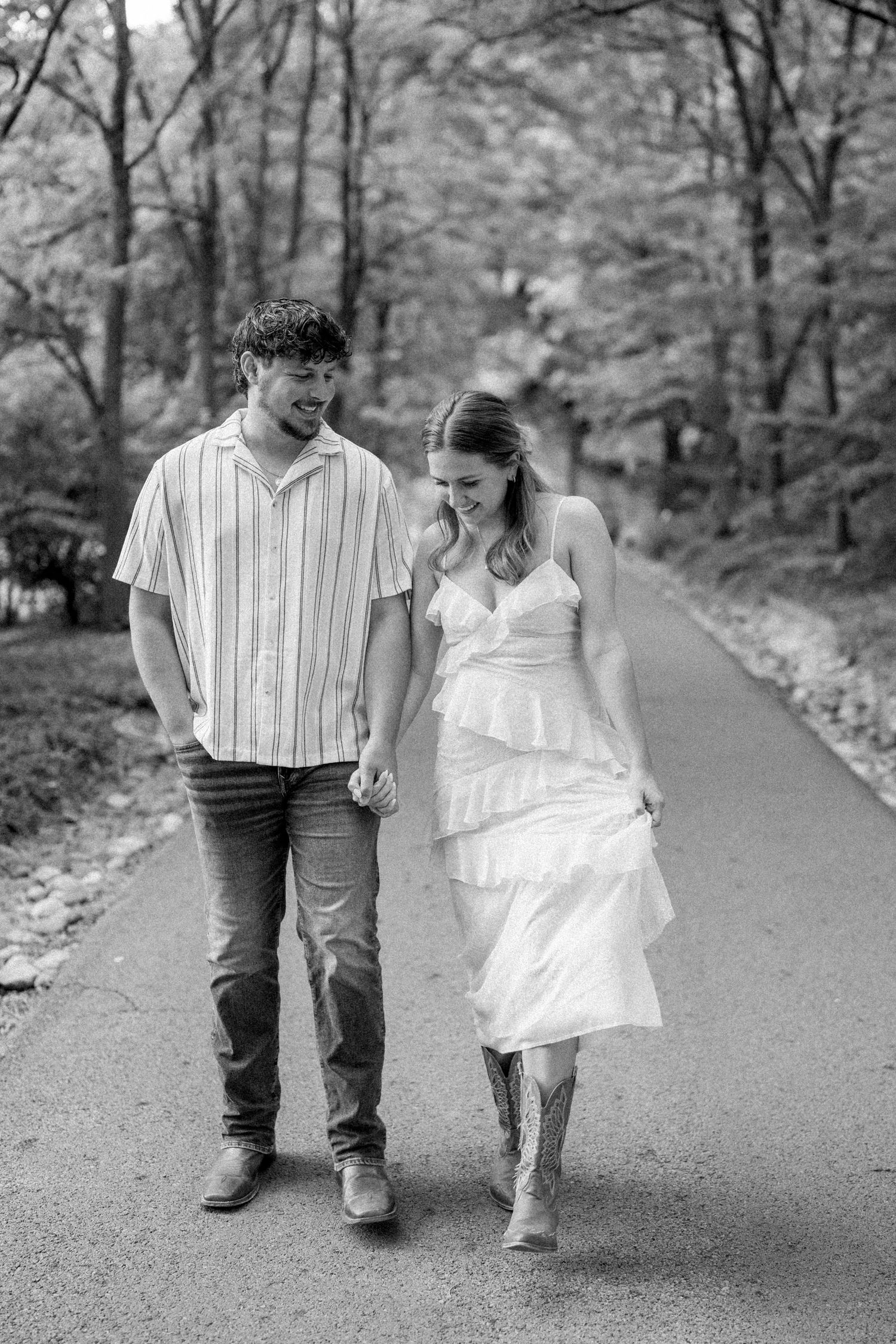 Black and white photo of a couple holding hands and walking down a tree-lined path, woman in white dress and cowboy boots.