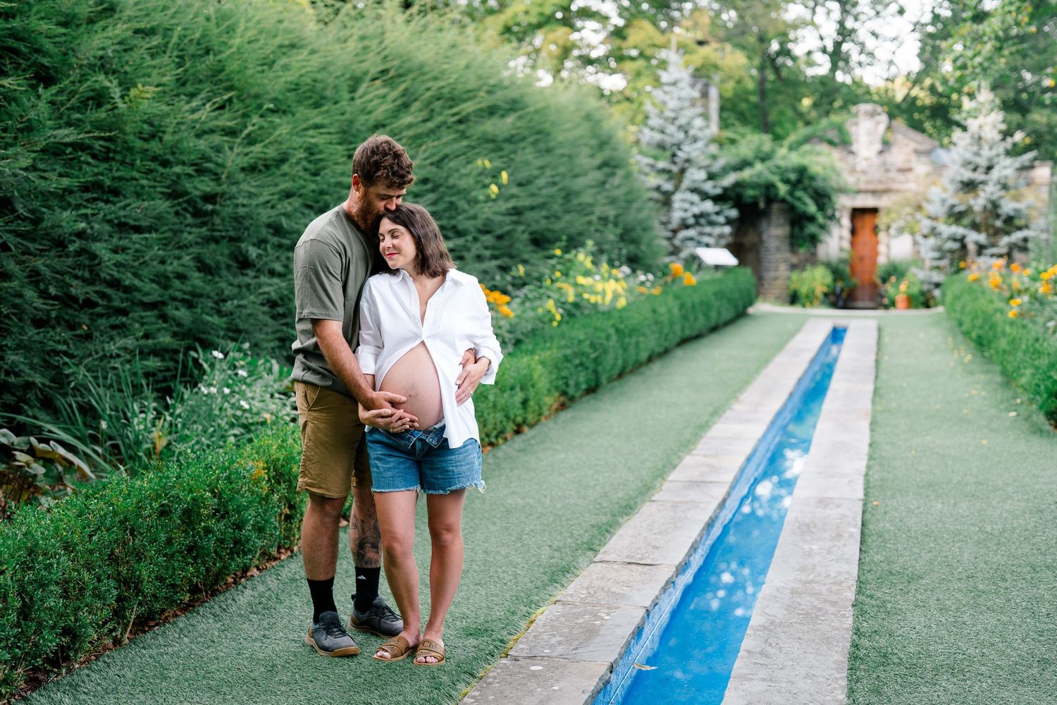 Couple embracing in a lush garden, pregnant woman in denim shorts with partner holding her belly outdoors.