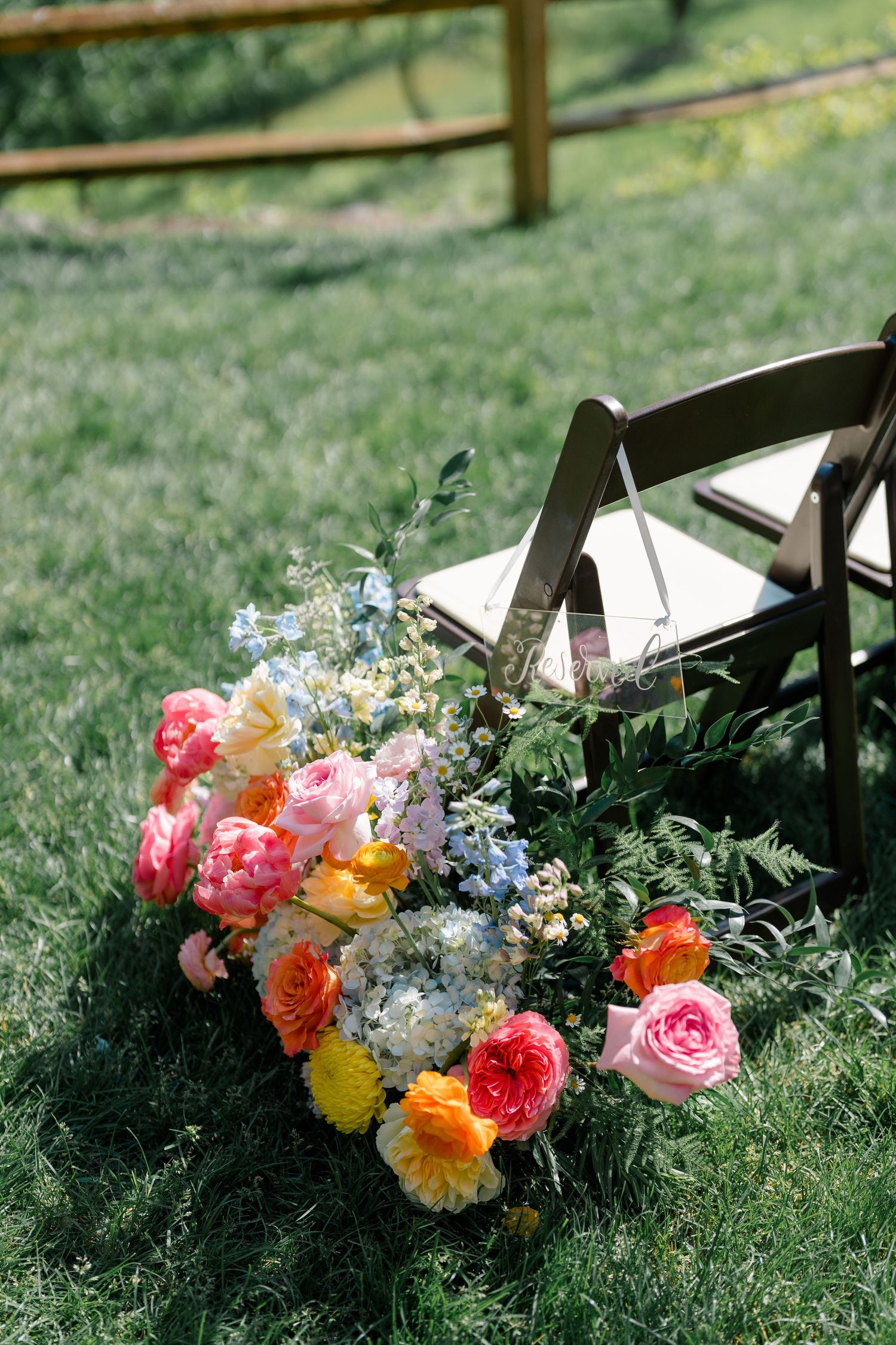 Vibrant pink, orange and blue floral arrangement rests on grass beside a wooden chair at an outdoor wedding.