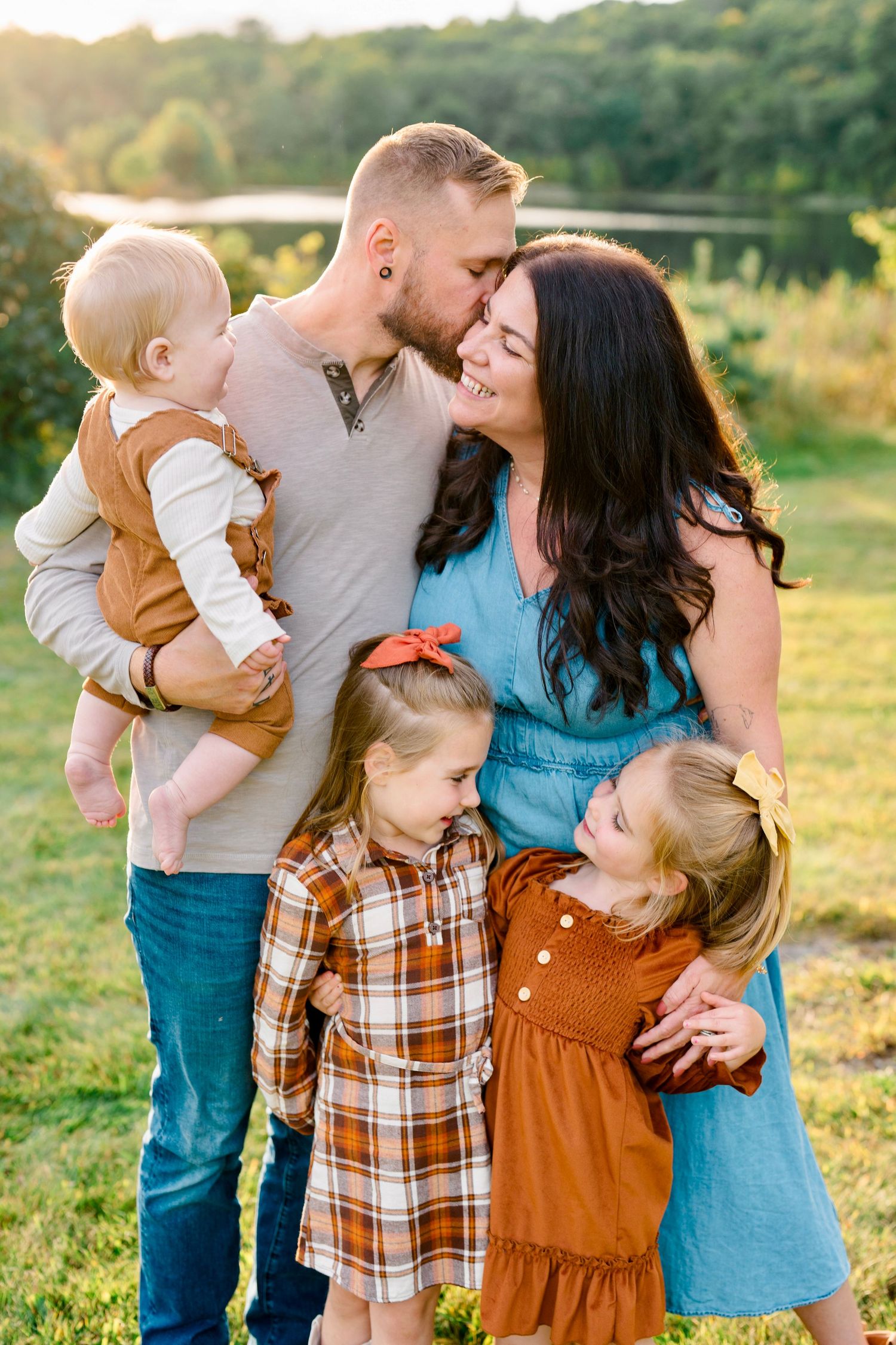A happy family of five poses outdoors at golden hour, with parents embracing three young daughters in autumn attire.