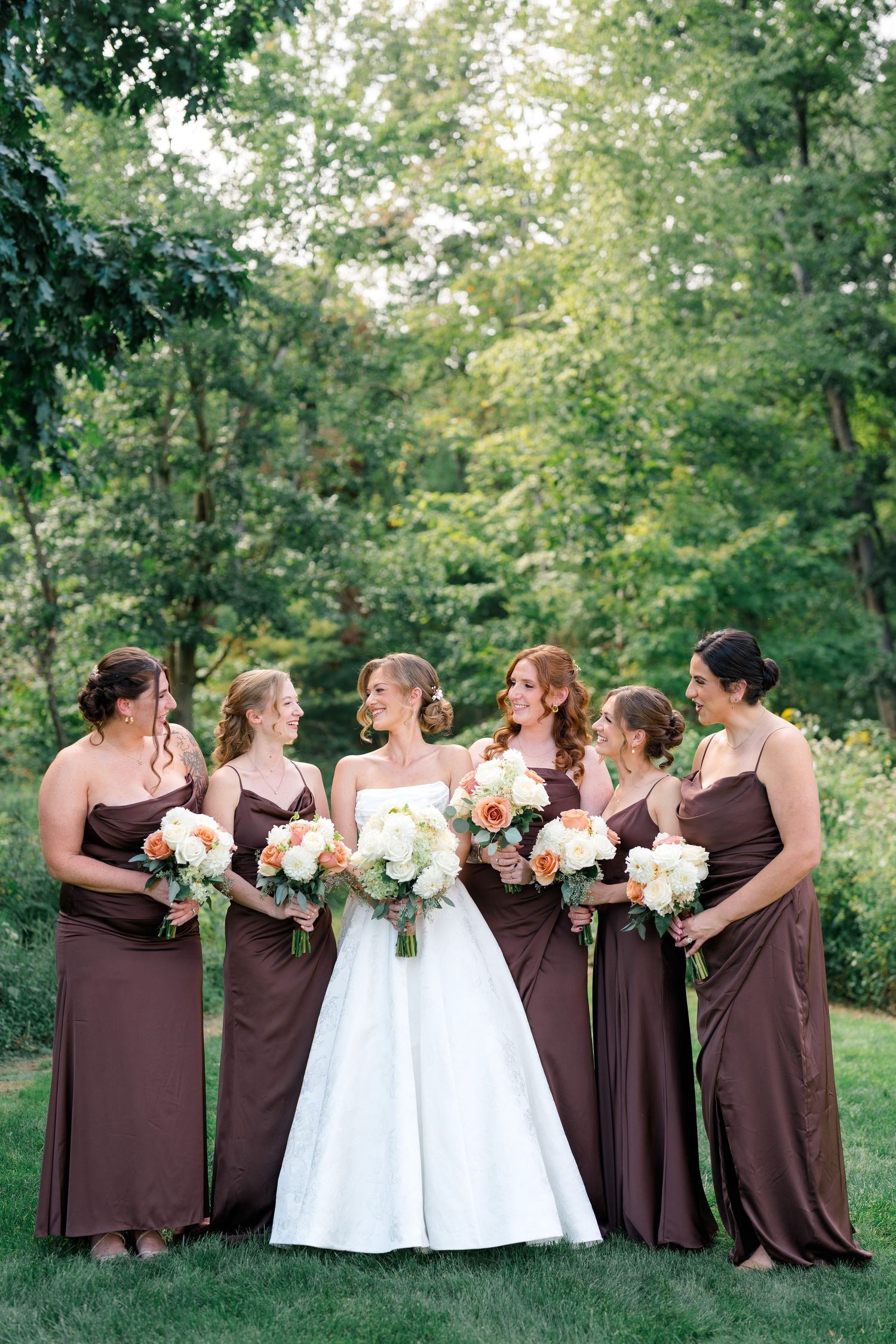 Bride in white strapless gown with five bridesmaids in brown dresses holding white and peach bouquets outdoors.