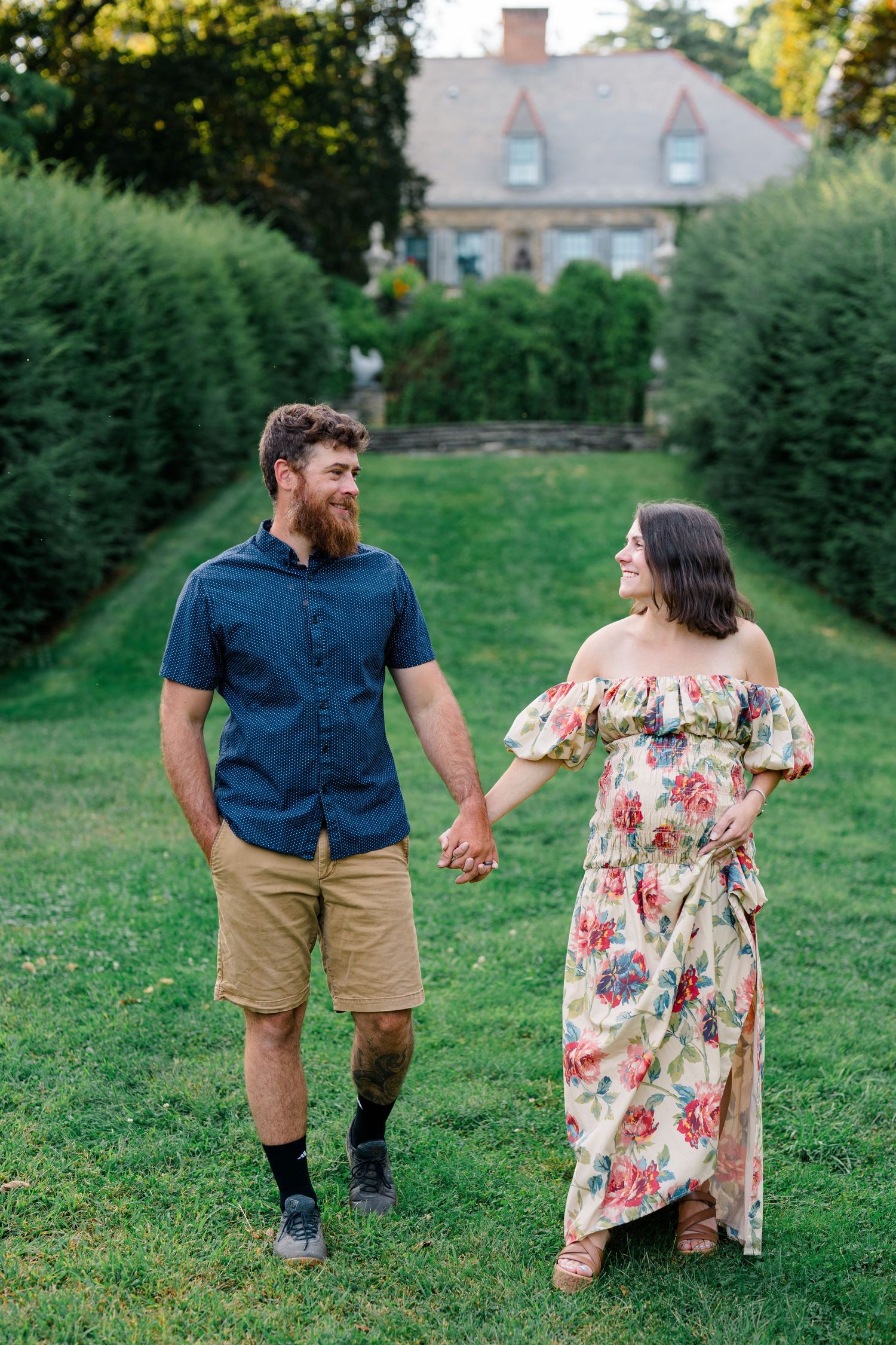 Smiling couple holding hands on green lawn with hedges and historic building, man in blue shirt and woman in floral dress.