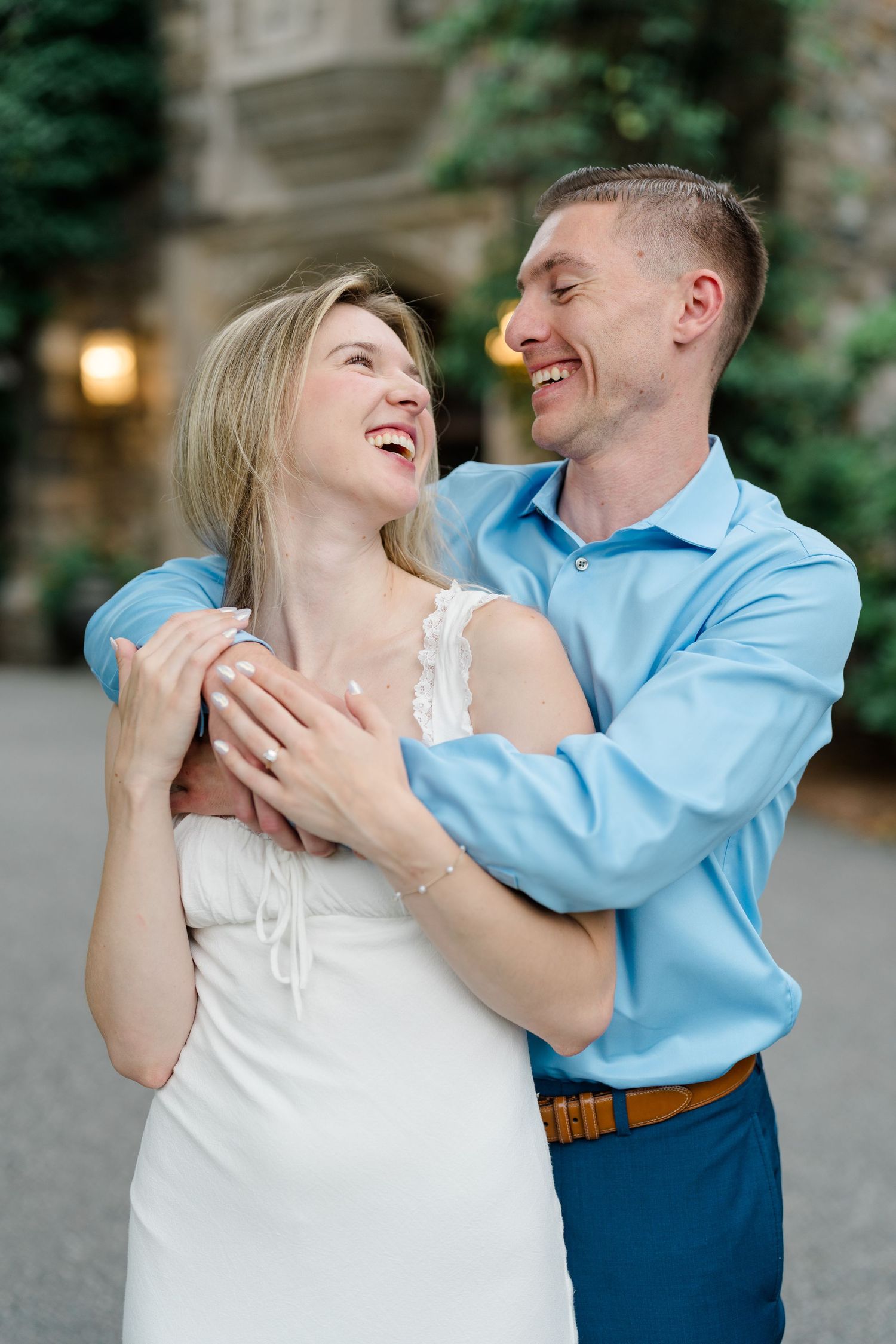 Happy couple embracing and laughing together outdoors, man in blue shirt hugging woman in white dress near stone building.