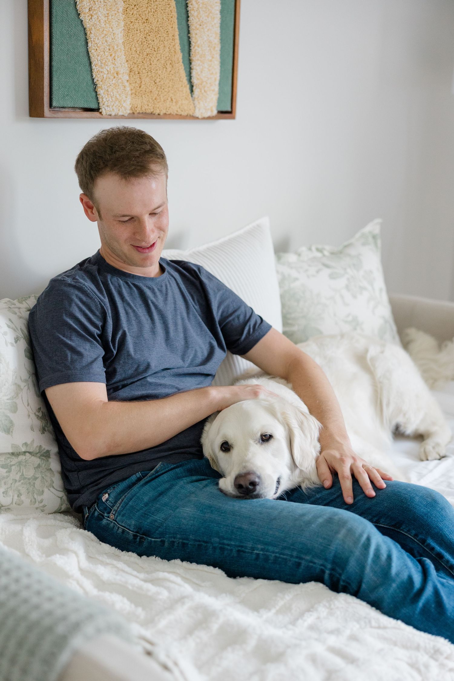 A couple relaxes with their white dog on a bed with fluffy white pillows and a teal wall artwork in the background.