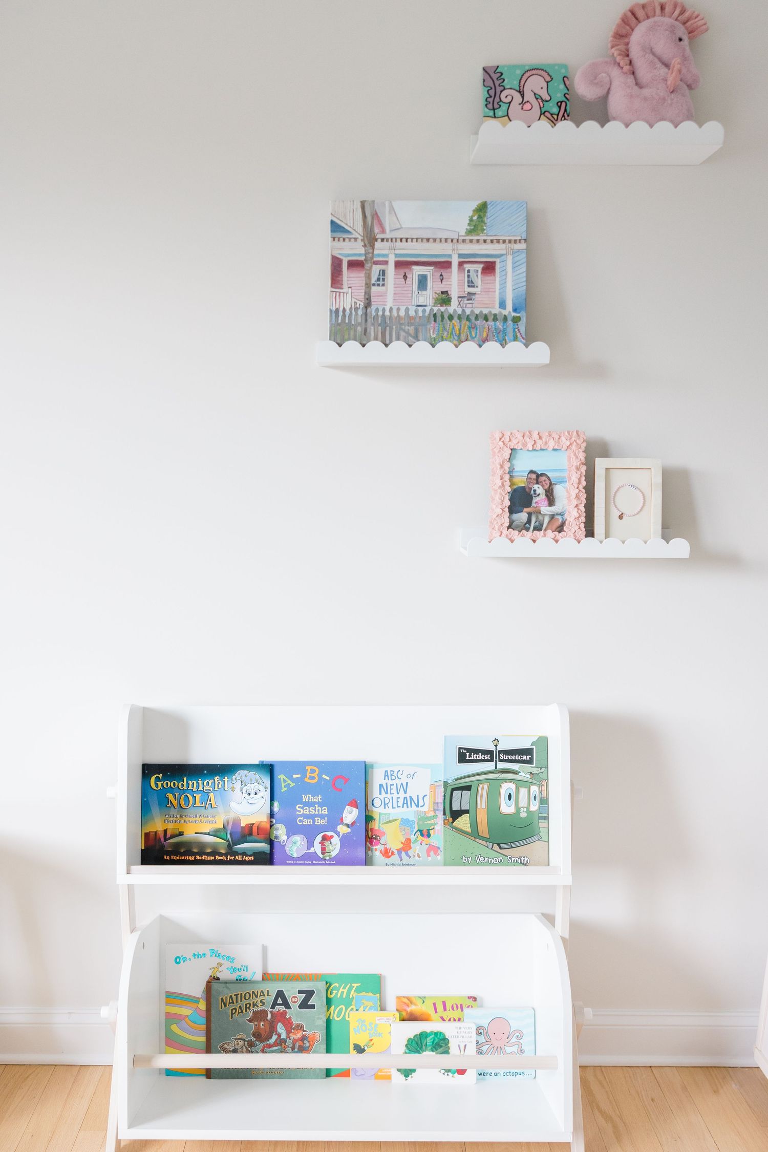 White nursery wall with floating shelves displaying children's books, toys and decor in soft pink and white colors.