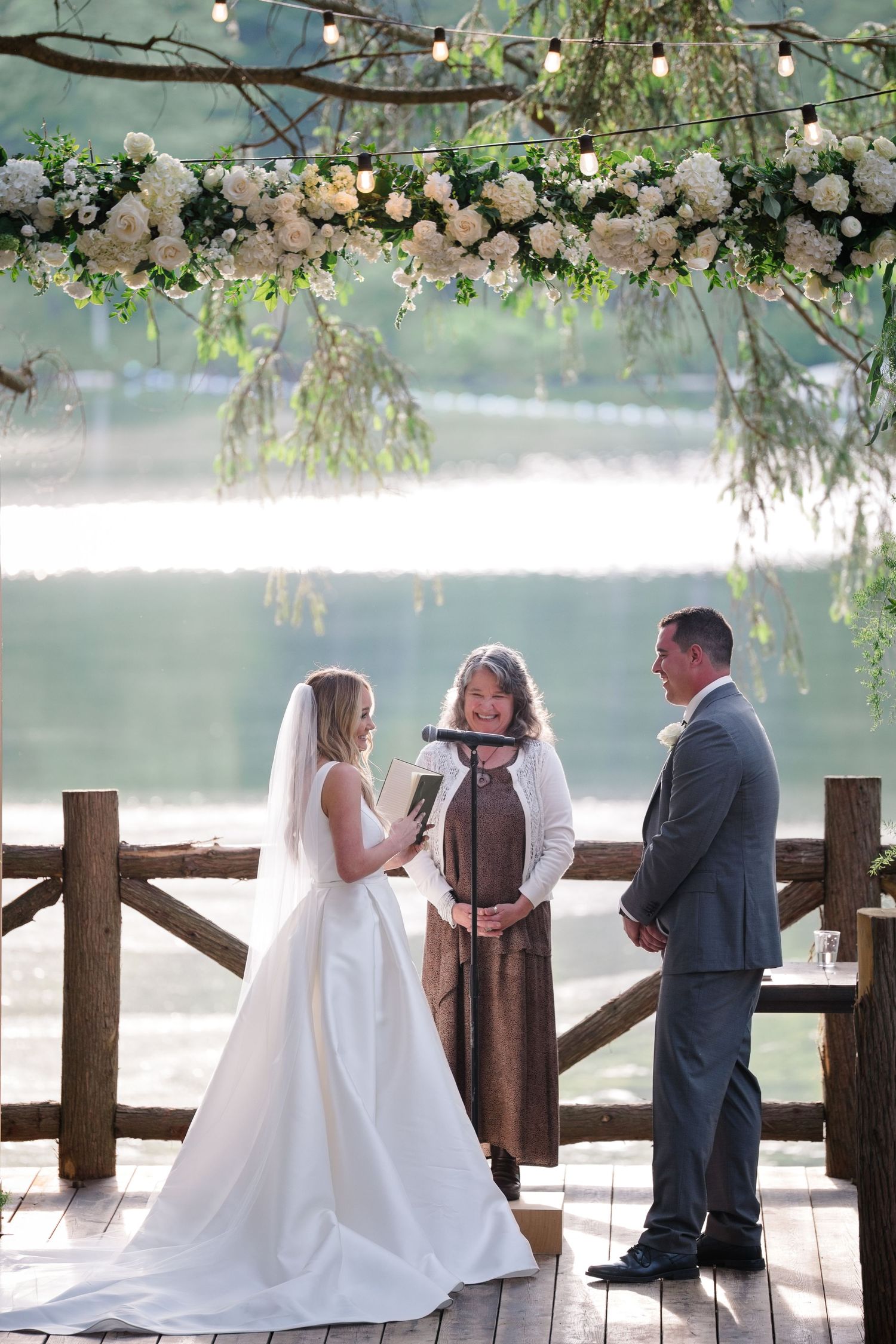 A romantic lakeside wedding ceremony takes place on a wooden dock decorated with white floral arrangements and draped greenery.