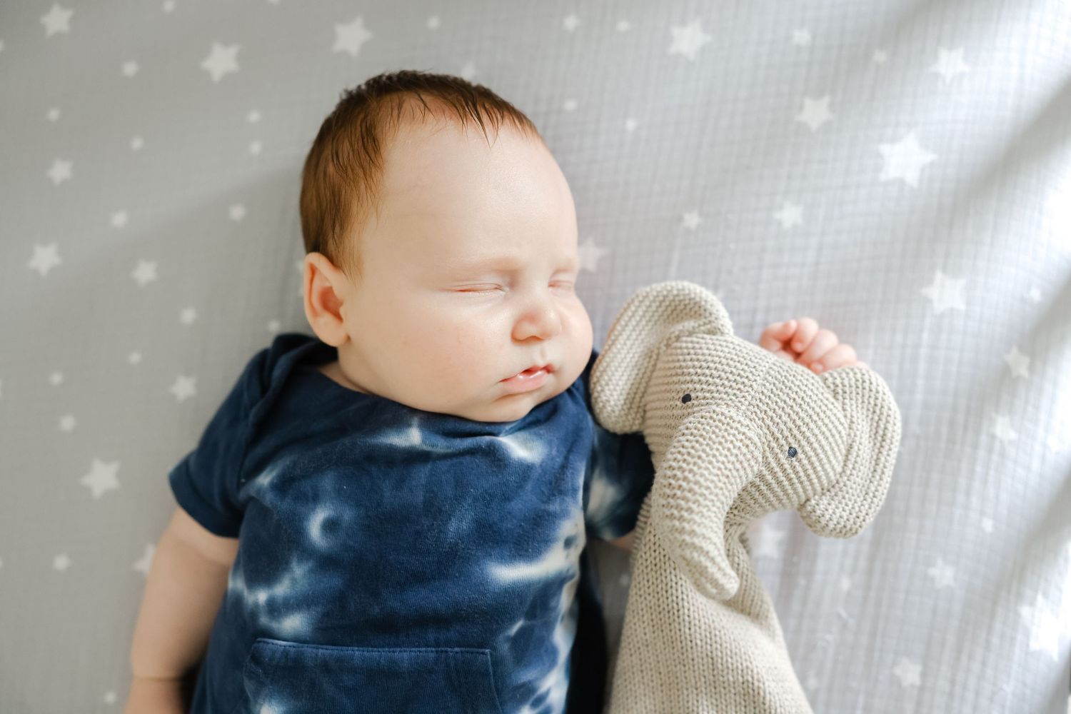 Sleeping baby in navy blue tie-dye shirt holds beige knitted stuffed animal on starry white background.