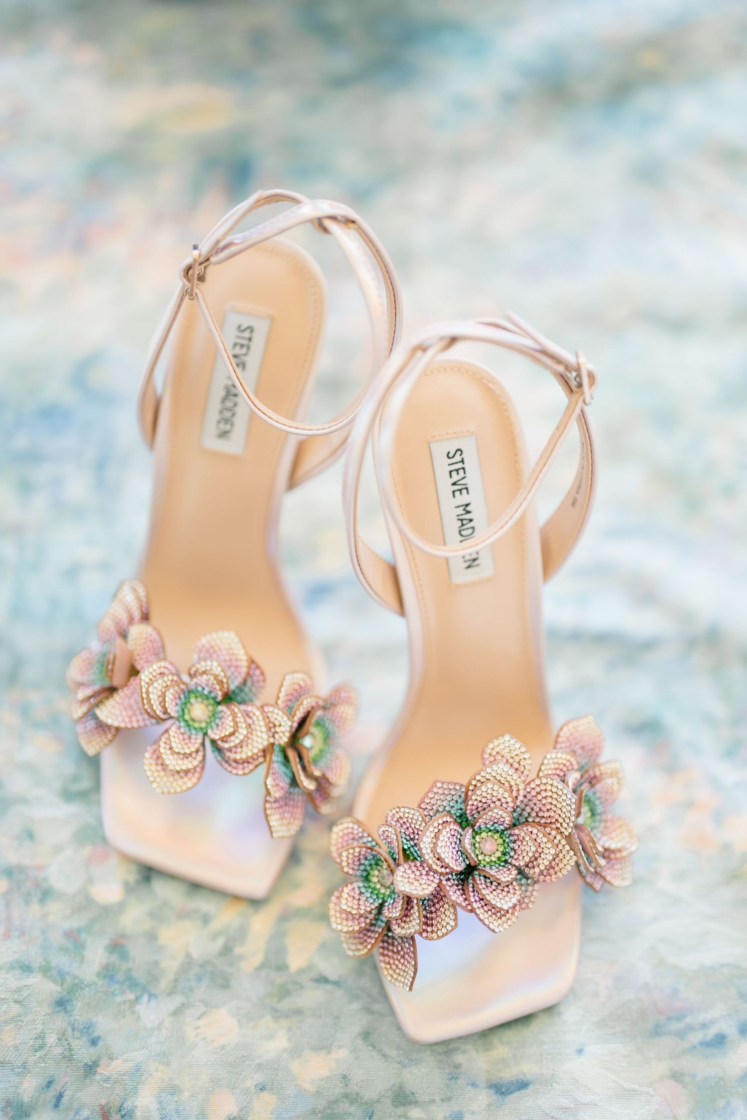 Elegant blush pink bridal shoes with crystal flower embellishments on a watercolor textured background.