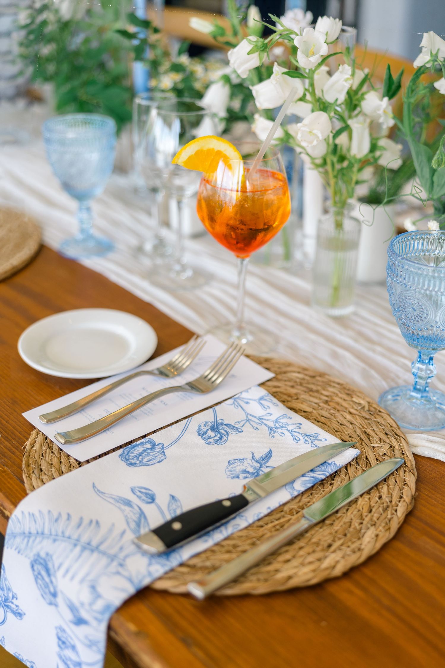 Elegant wedding place setting with blue floral napkin, wicker charger, and Aperol Spritz cocktail.