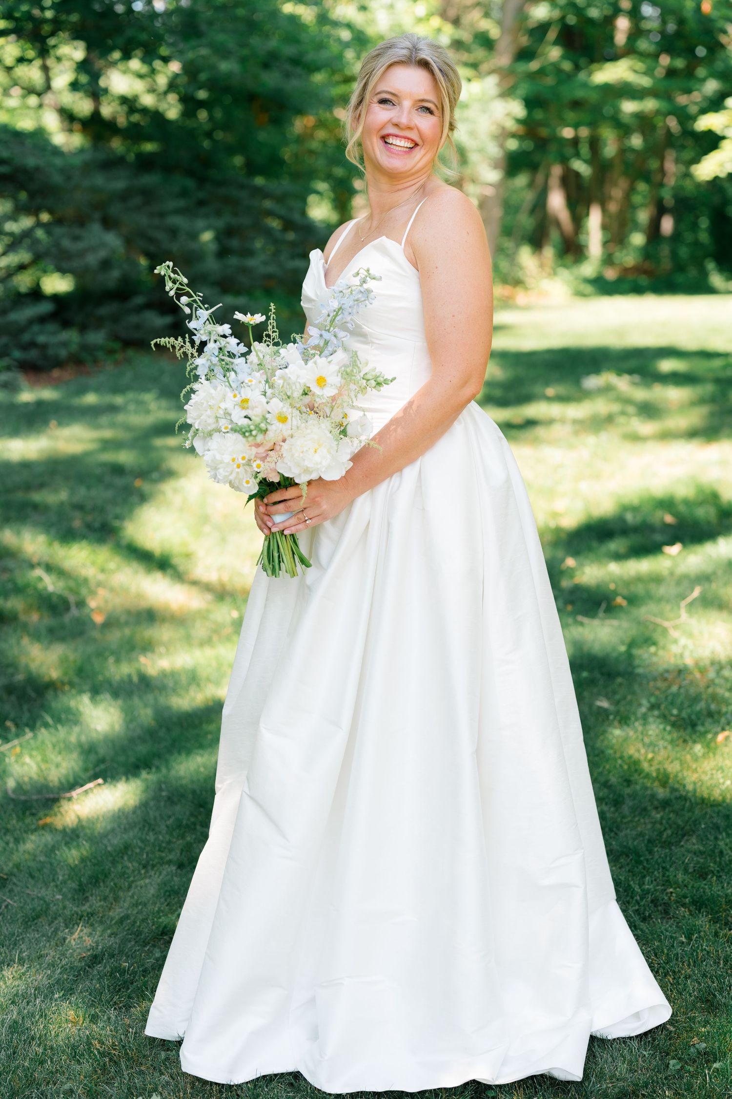 Smiling bride in white spaghetti-strap wedding gown holds white daisy bouquet outdoors in green garden setting.
