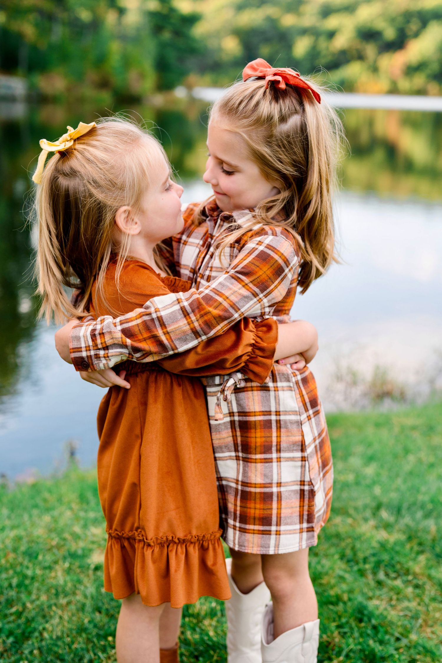 Two young girls in autumn dresses hugging near a lake, wearing hair bows in fall colors.