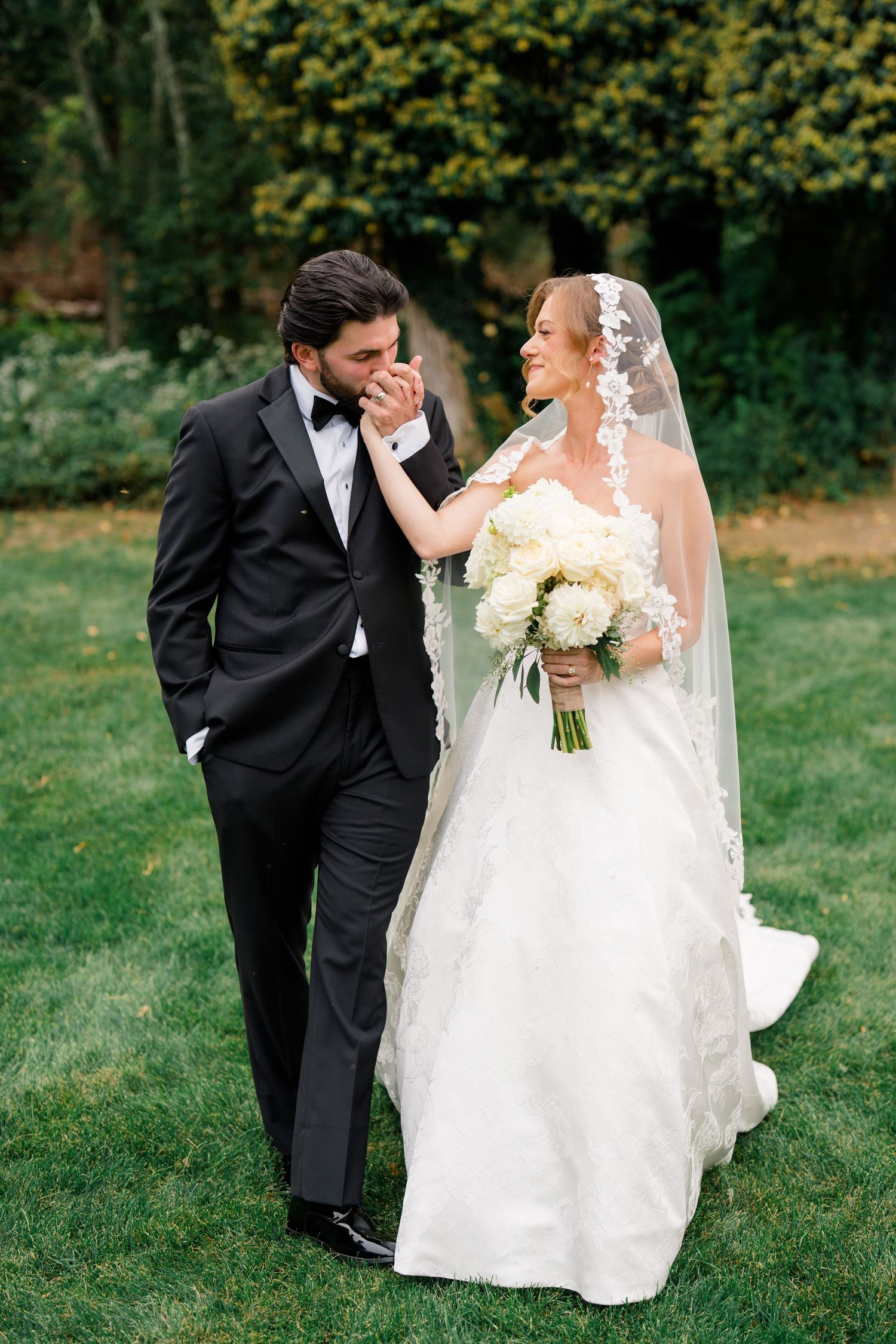 Groom in black tuxedo kisses bride's hand as she smiles holding white bouquet on green lawn.