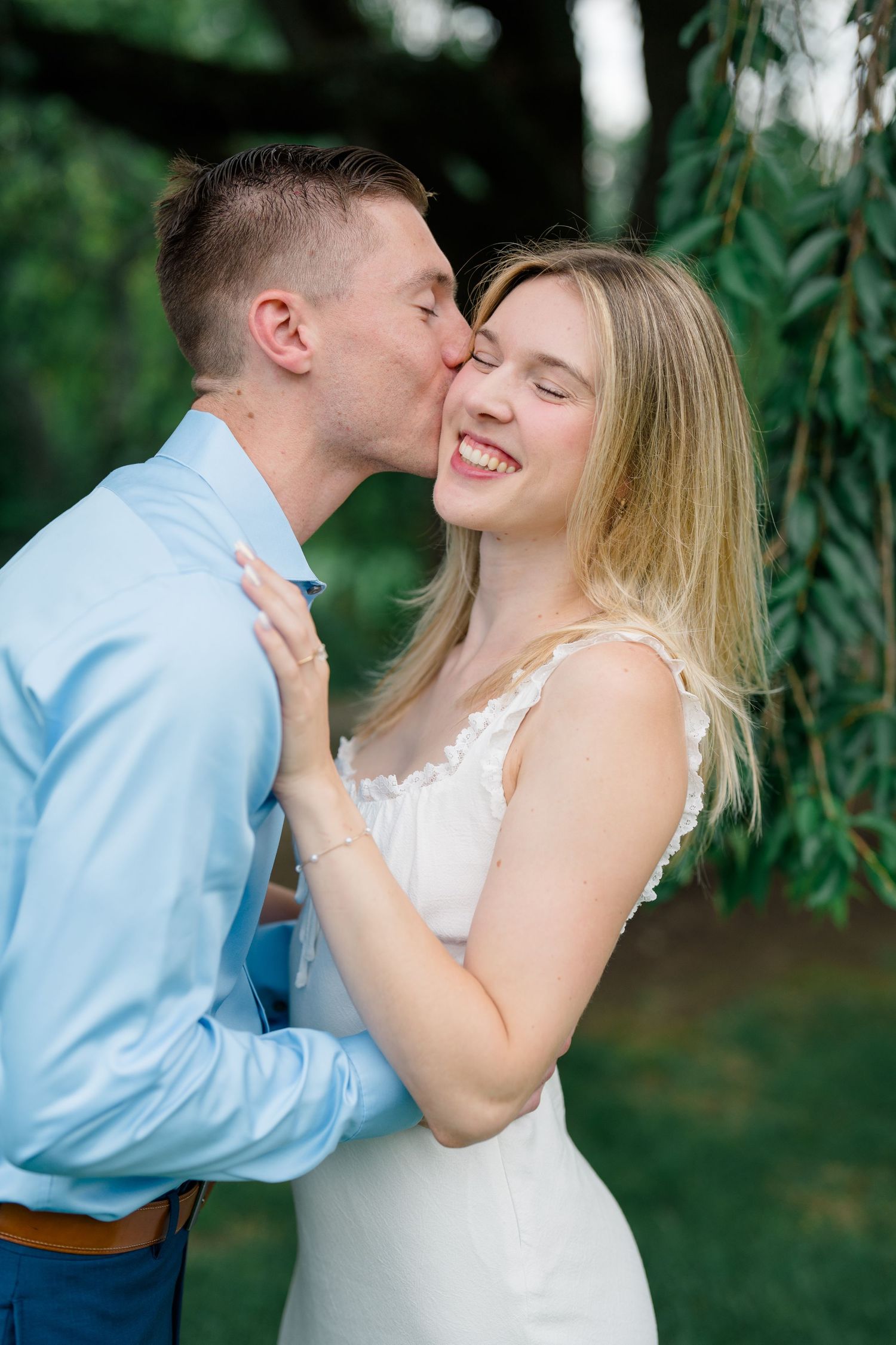 A smiling bride in white dress receives a cheek kiss from groom in blue shirt outdoors near greenery.
