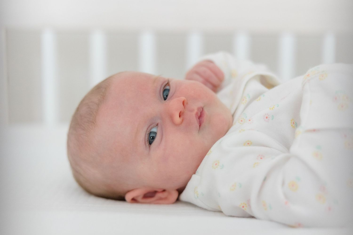 A baby wearing white clothing lies down and moves their head while looking up.
