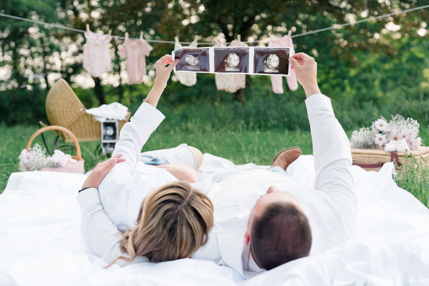 Two people lying on white blanket outdoors looking at ultrasound photos during pregnancy shoot.