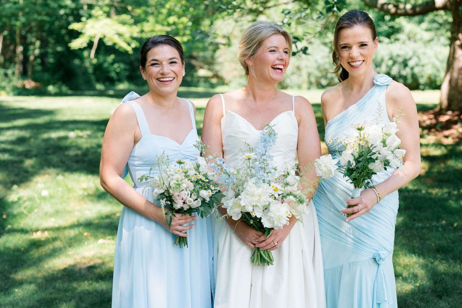 Bride and two bridesmaids in light blue dresses holding white floral bouquets, smiling outdoors in a lush green garden.