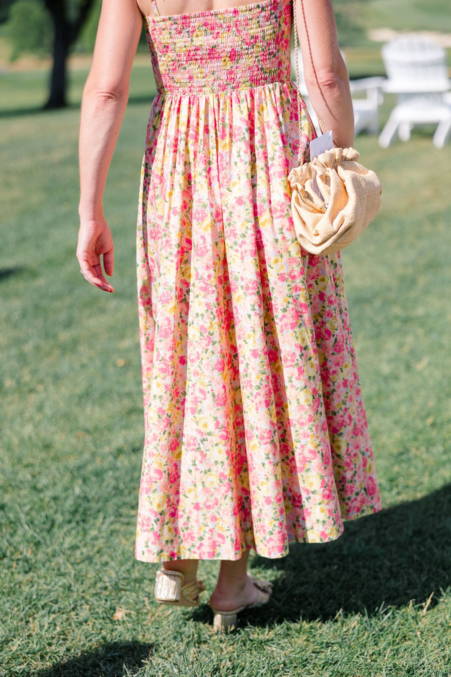 Woman wearing pink floral smocked midi dress with straw bag and nude heels walking on green lawn.