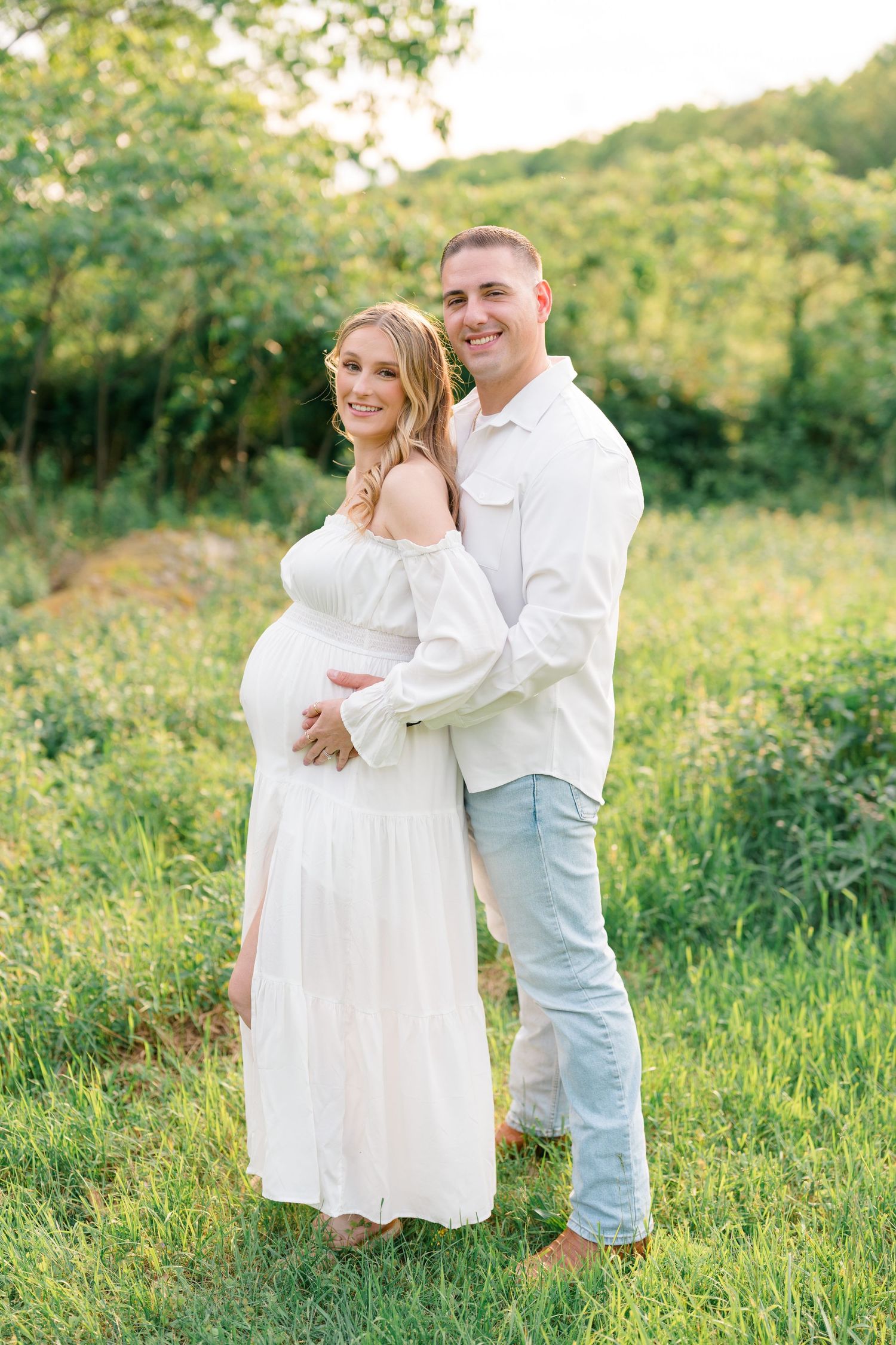 Romantic maternity photo of couple embracing in a sunlit meadow wearing coordinated white and denim outfits.