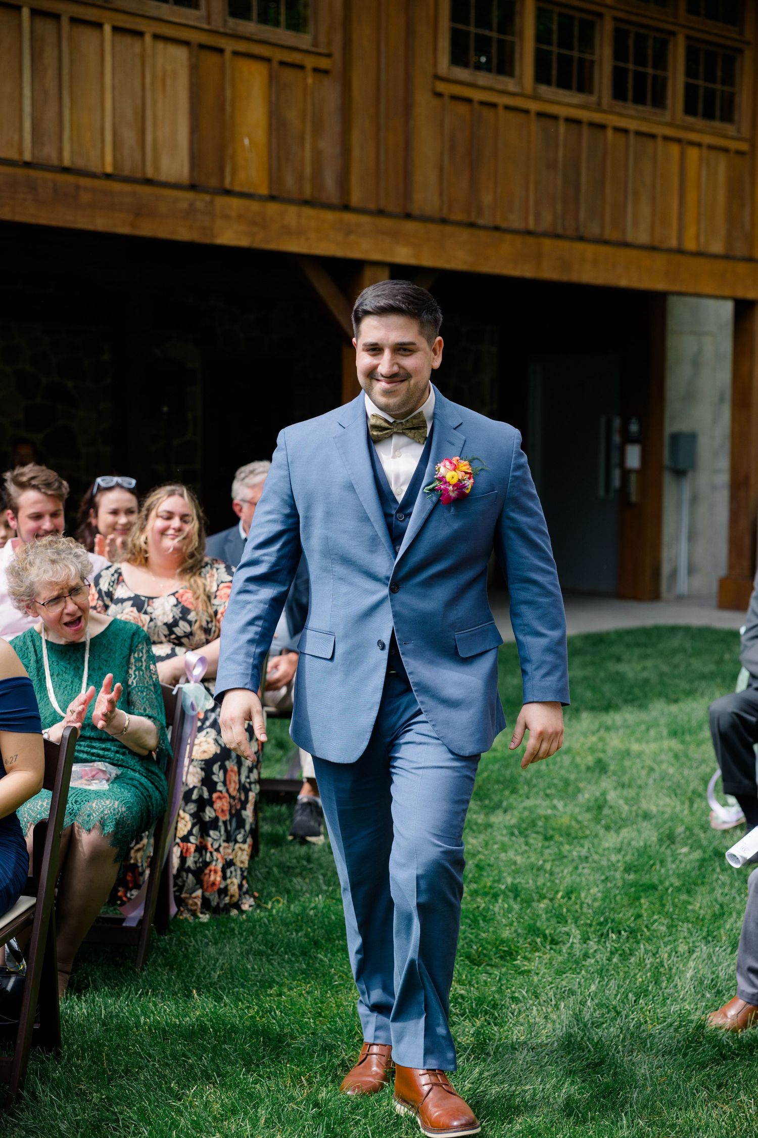 Groom in blue suit and brown shoes walks down grassy aisle at outdoor wedding ceremony.