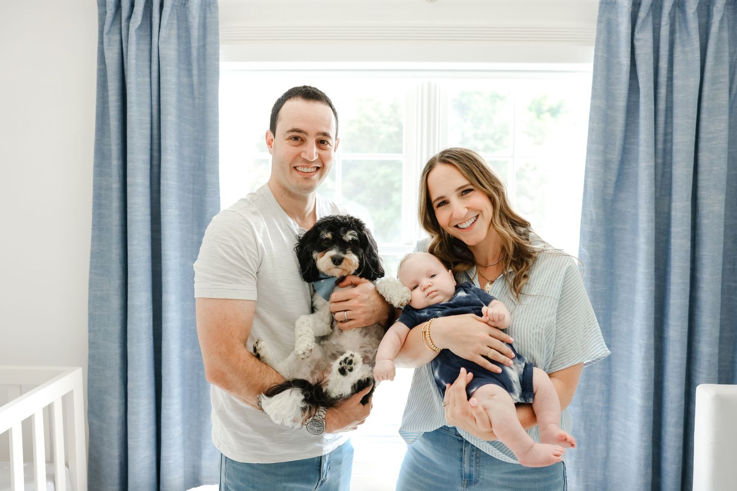People in gray and white casual clothing stand in a bright room with blue curtains while holding a baby and small dog.