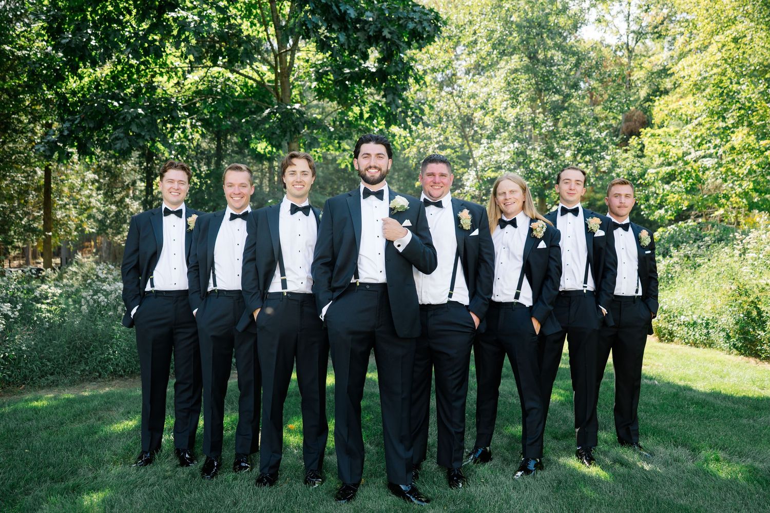 Seven groomsmen in black tuxedos with bow ties pose outdoors on green grass surrounded by lush trees at a wedding.