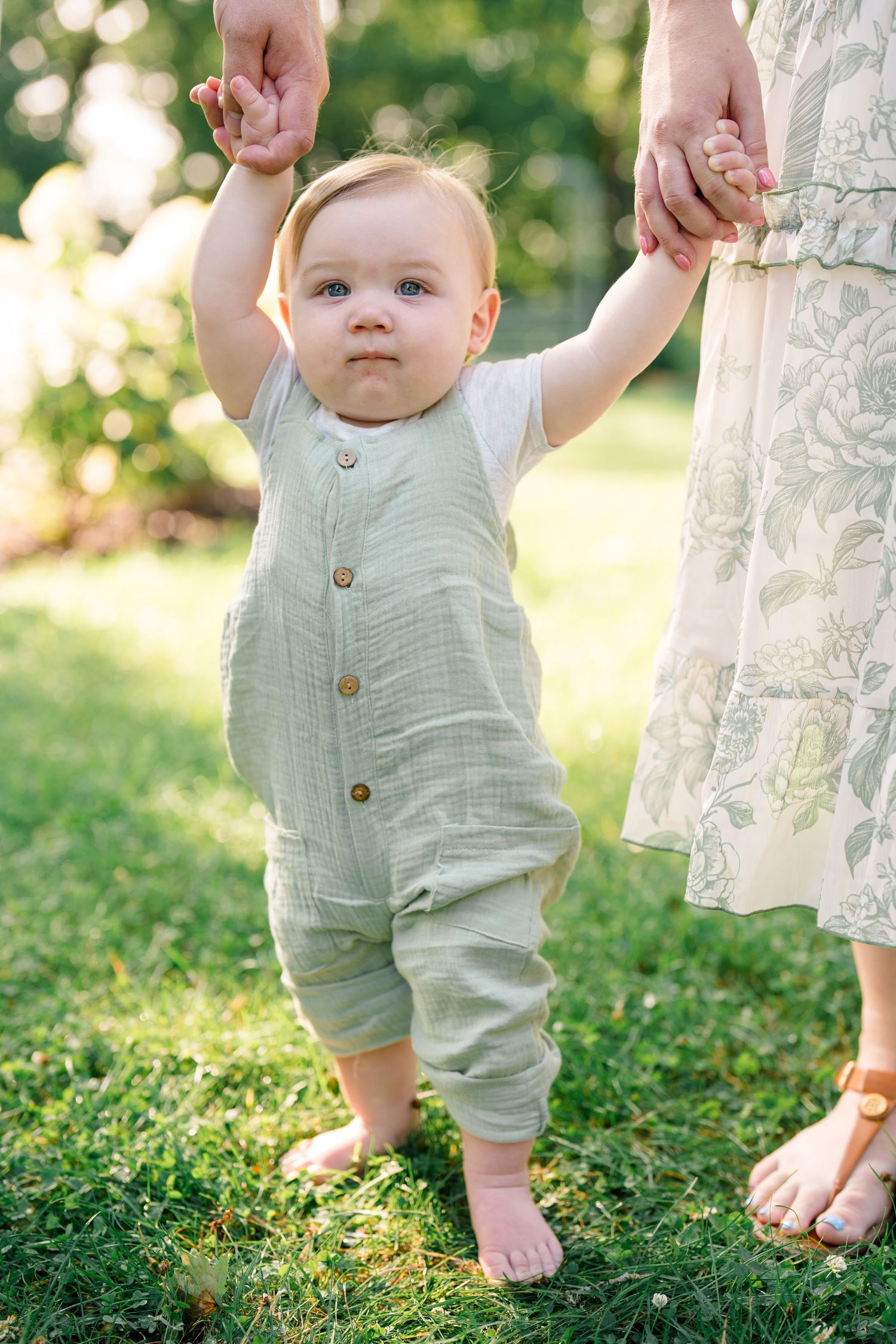 An adorable baby in a sage green linen romper takes first steps barefoot on grass, holding mothers hands outdoors.