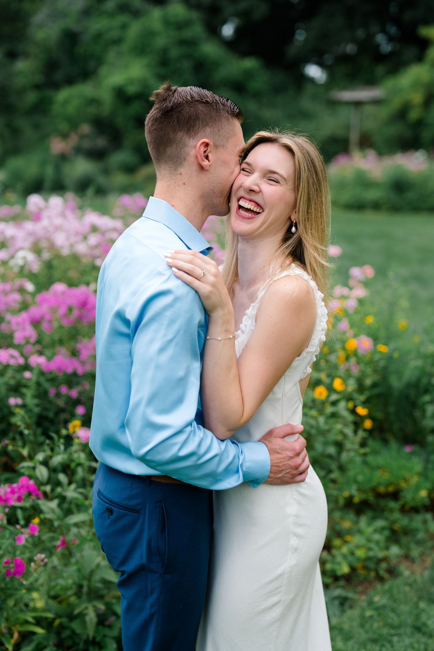 A laughing couple embraces in a colorful flower garden, with the man whispering in the woman's ear.