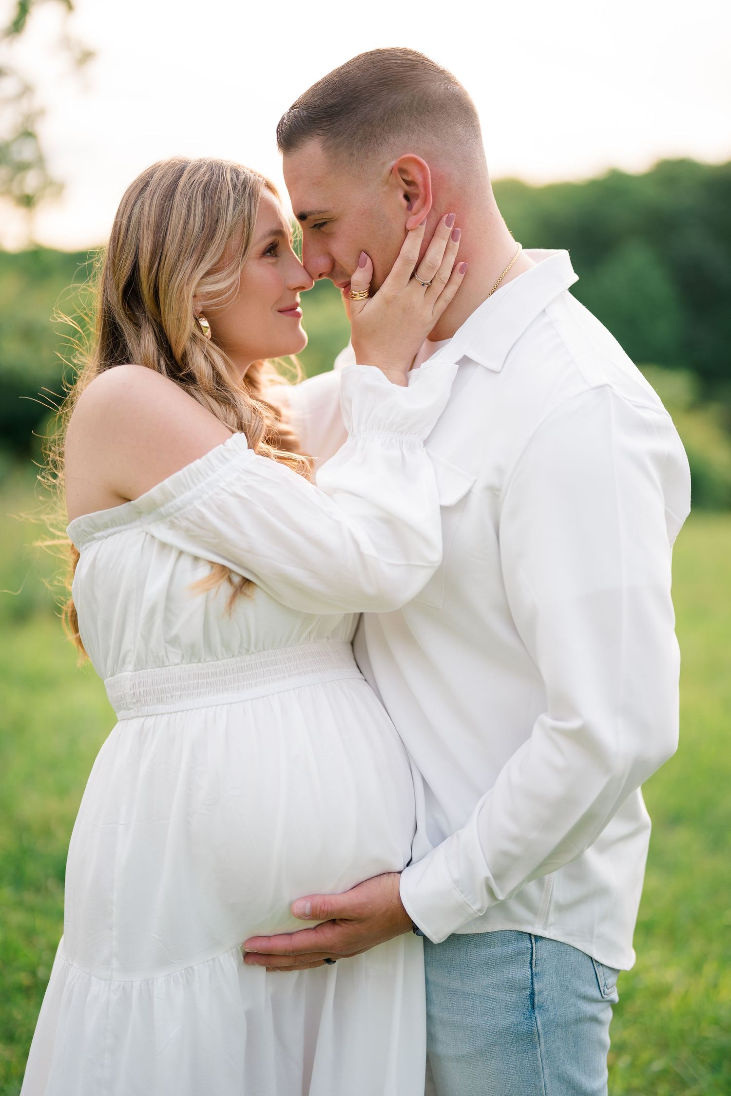 Tender maternity photography captures couple's embrace in white outfits against green backdrop.