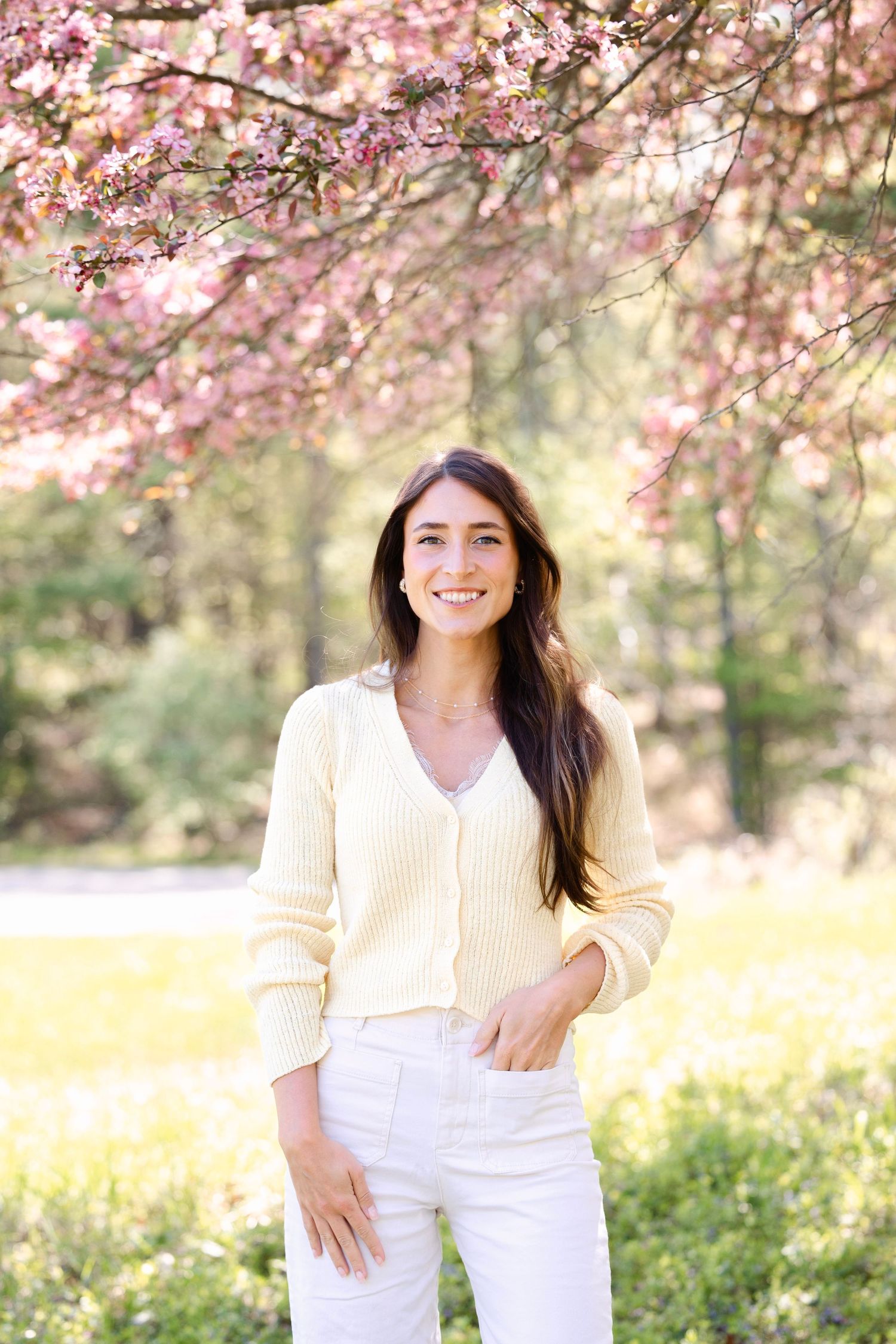 Young woman in yellow sweater and white jeans smiling under pink cherry blossom tree in sunny park.