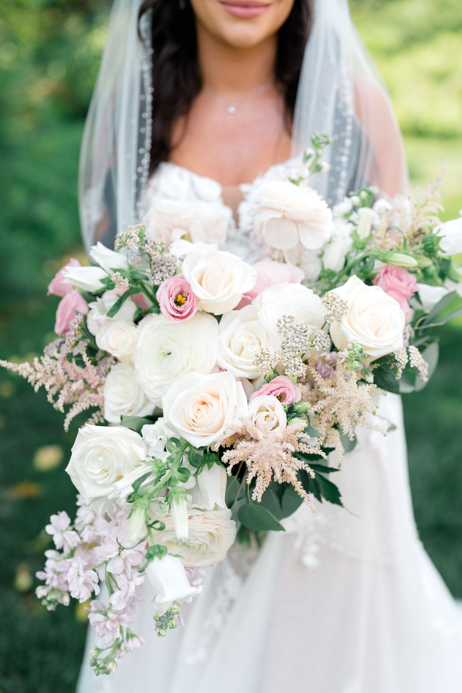 Elegant white and pink wedding bouquet with roses and delicate greenery held against a natural outdoor backdrop.