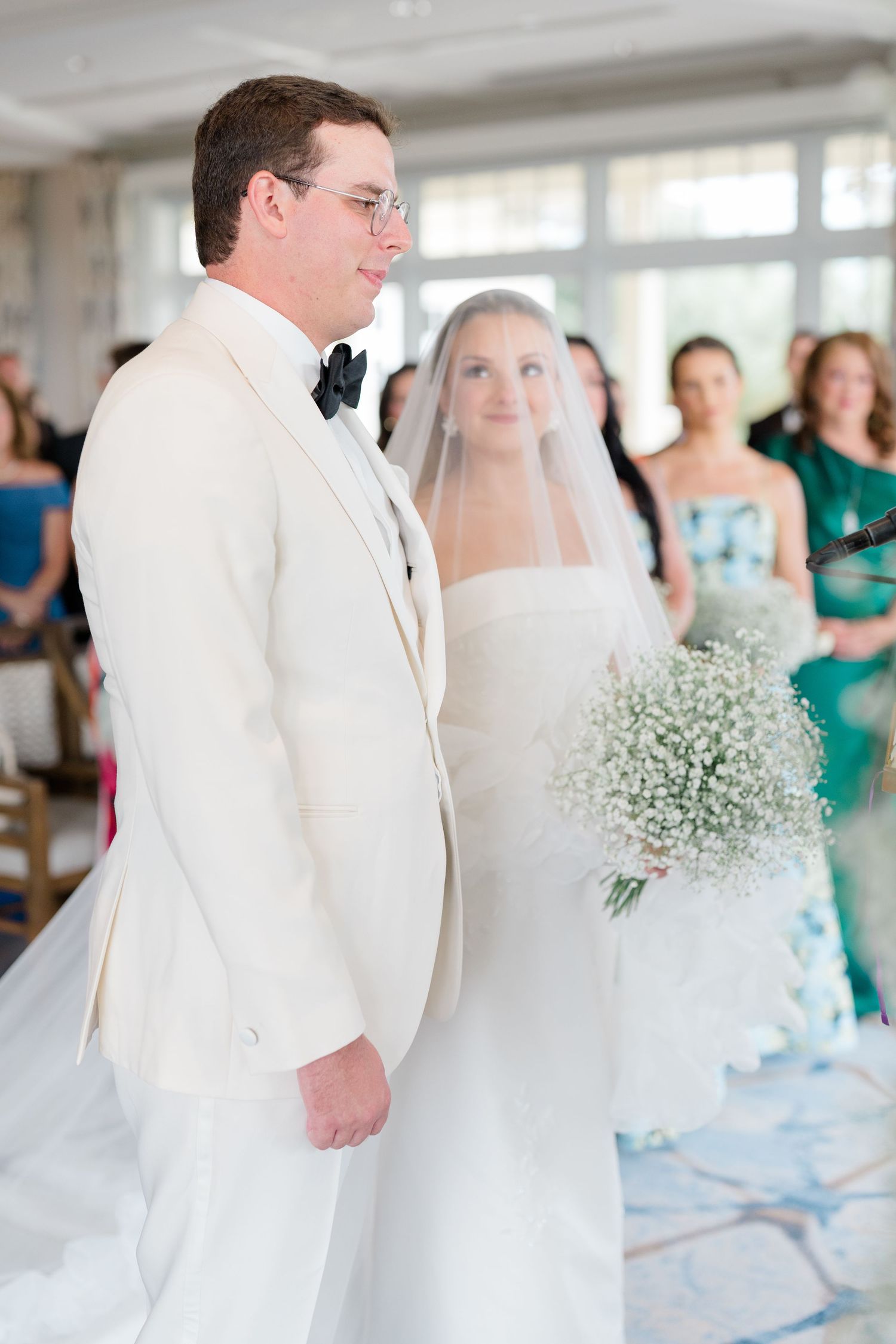 Wedding couple prepares to dance in white attire with bridesmaids in teal dresses and baby's breath bouquets at reception.