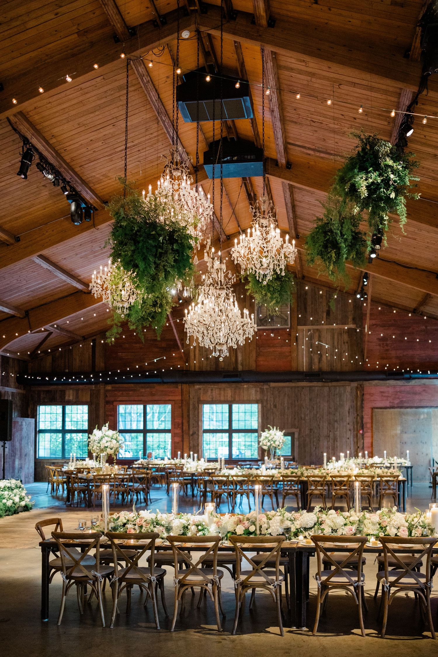 Elegant rustic wedding reception venue with wooden beams, crystal chandeliers, string lights and long dining tables.