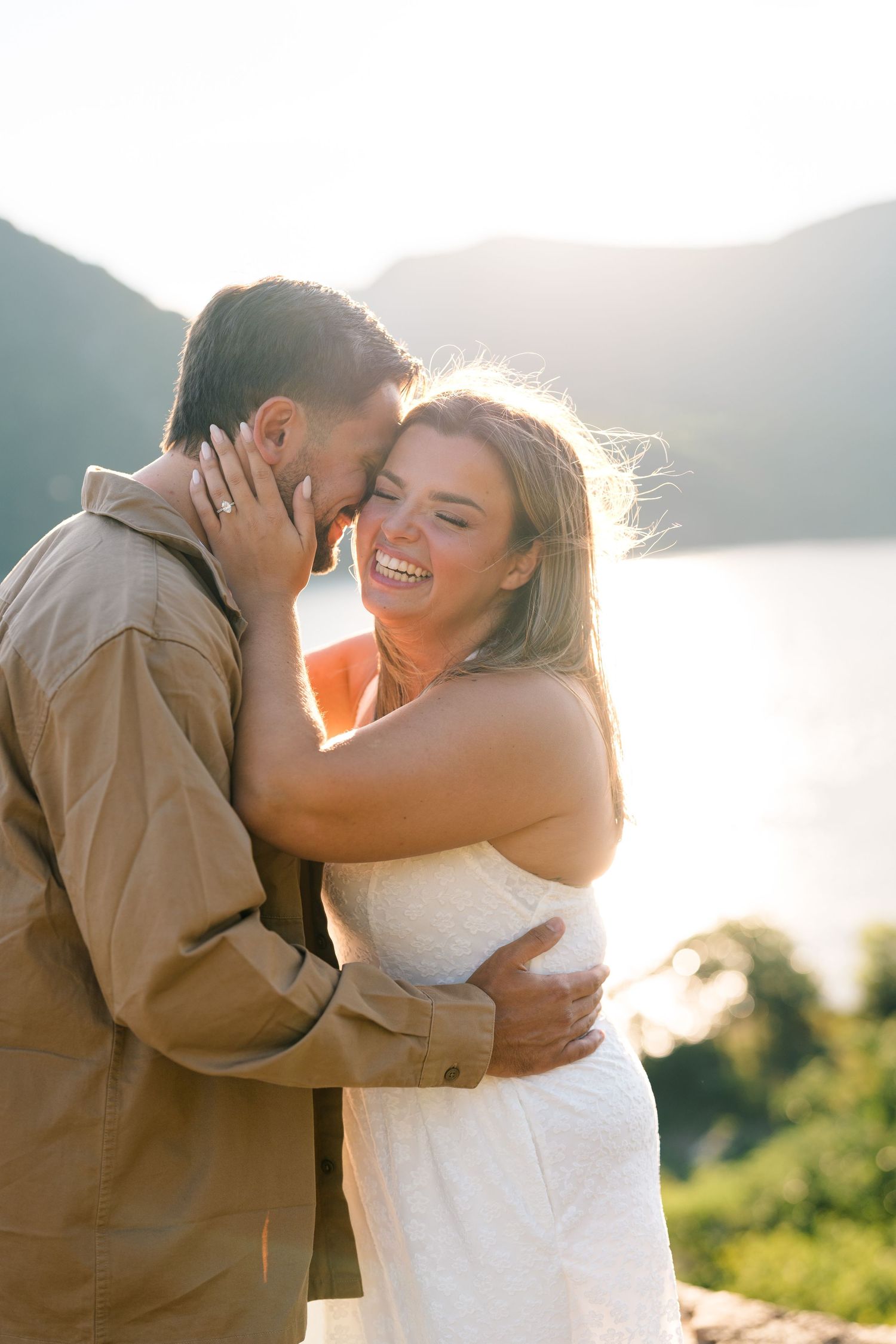 Happy couple embracing at sunset with mountain lake backdrop, woman in white dress smiling joyfully.