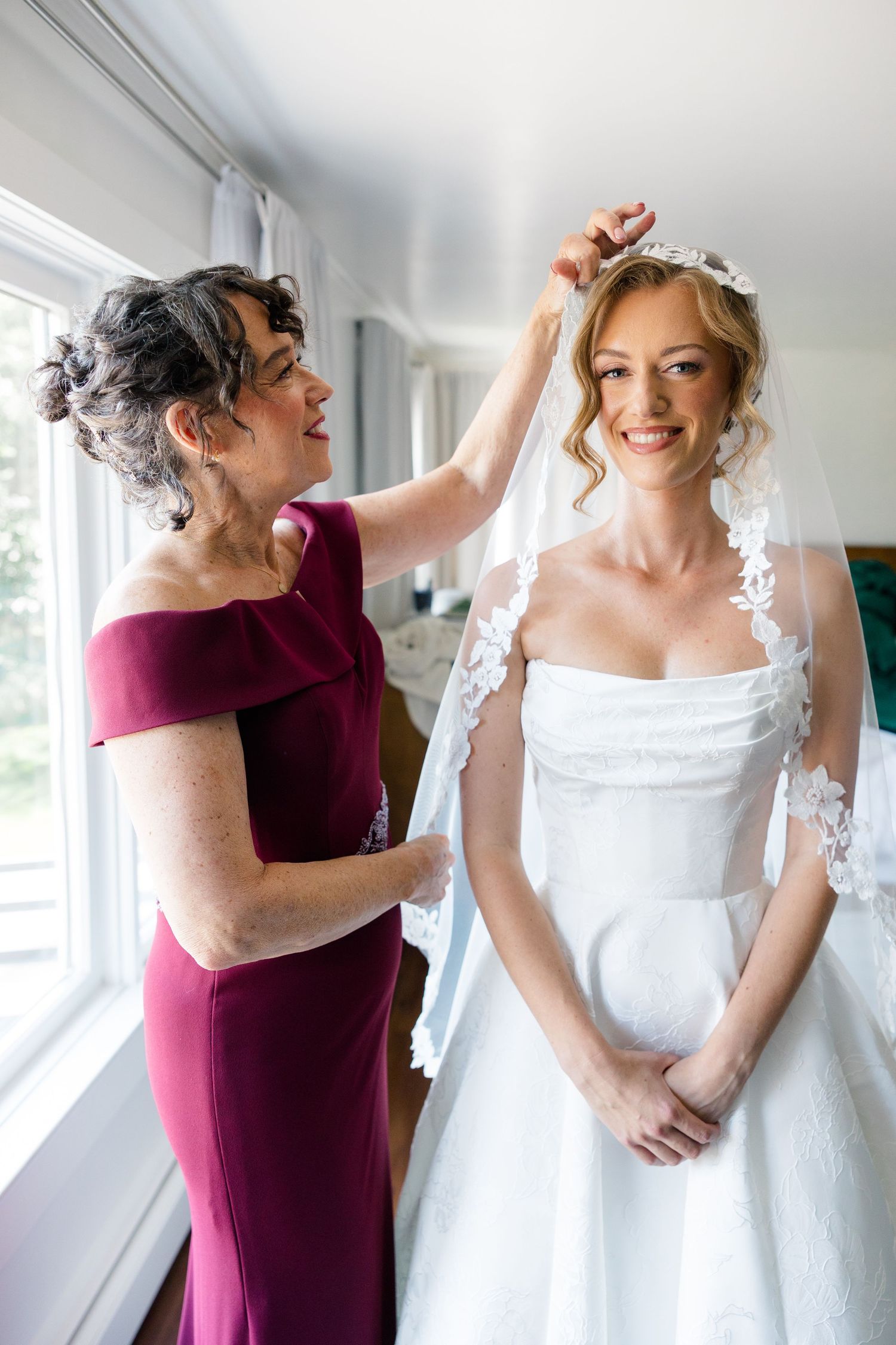 Mother of the bride adjusts lace veil on smiling bride in white strapless gown near bright window.