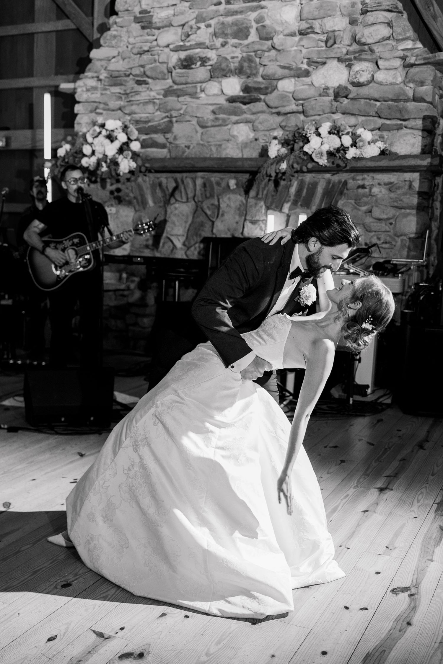 Black and white photo of bride and groom sharing a romantic dip kiss on dance floor with live band performing behind them.