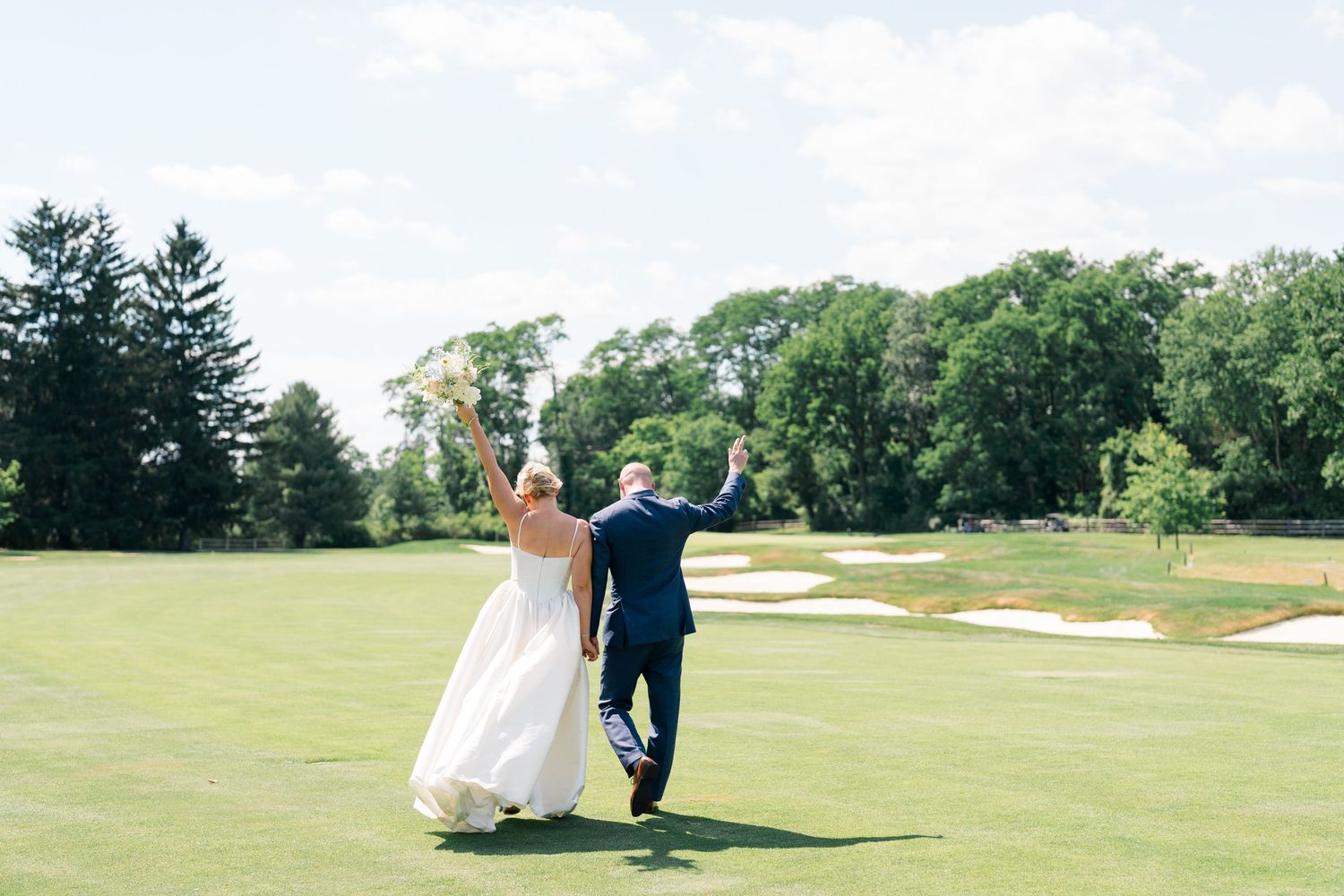 Bride and groom walking on golf course, arms raised in celebration, wearing wedding attire on sunny day.